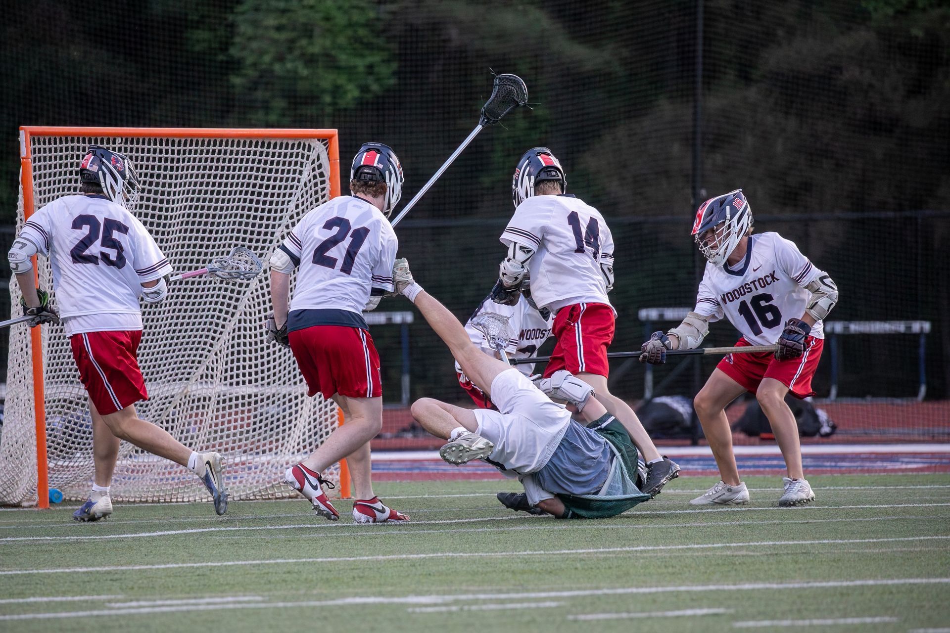 Lacrosse players in white and red clash near the goal as one player falls backward on the field.