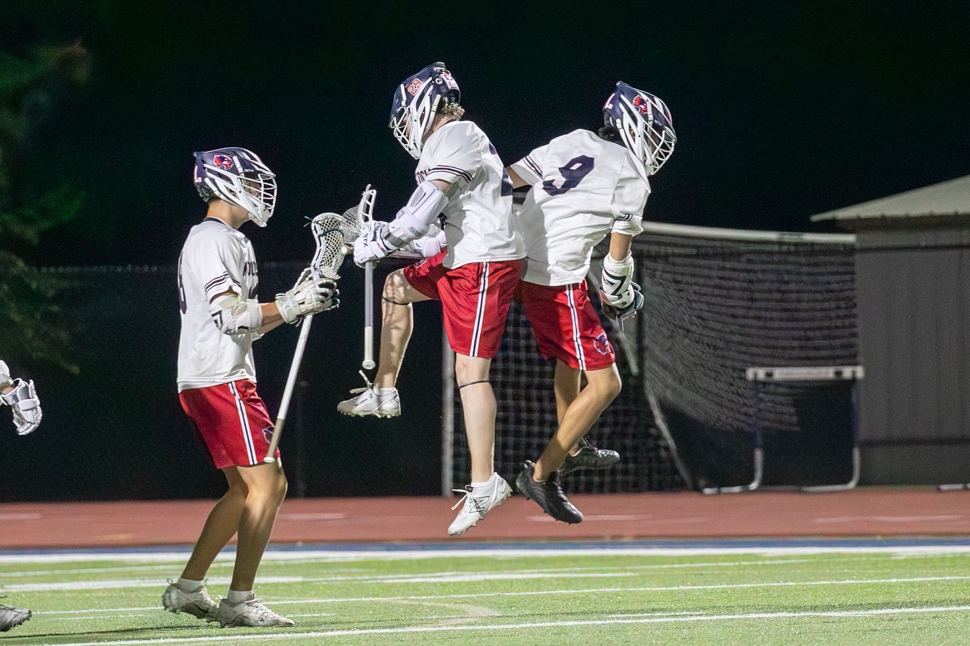 Three lacrosse players in white jerseys and red shorts jump celebrating on a field at night.