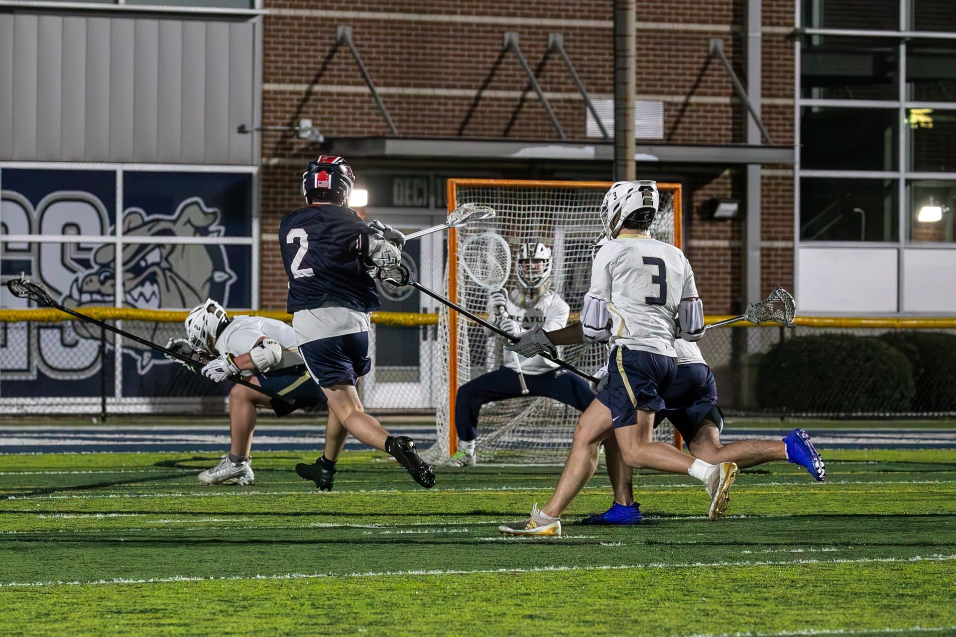 Lacrosse players in white and dark uniforms near the goal on a lit field at night