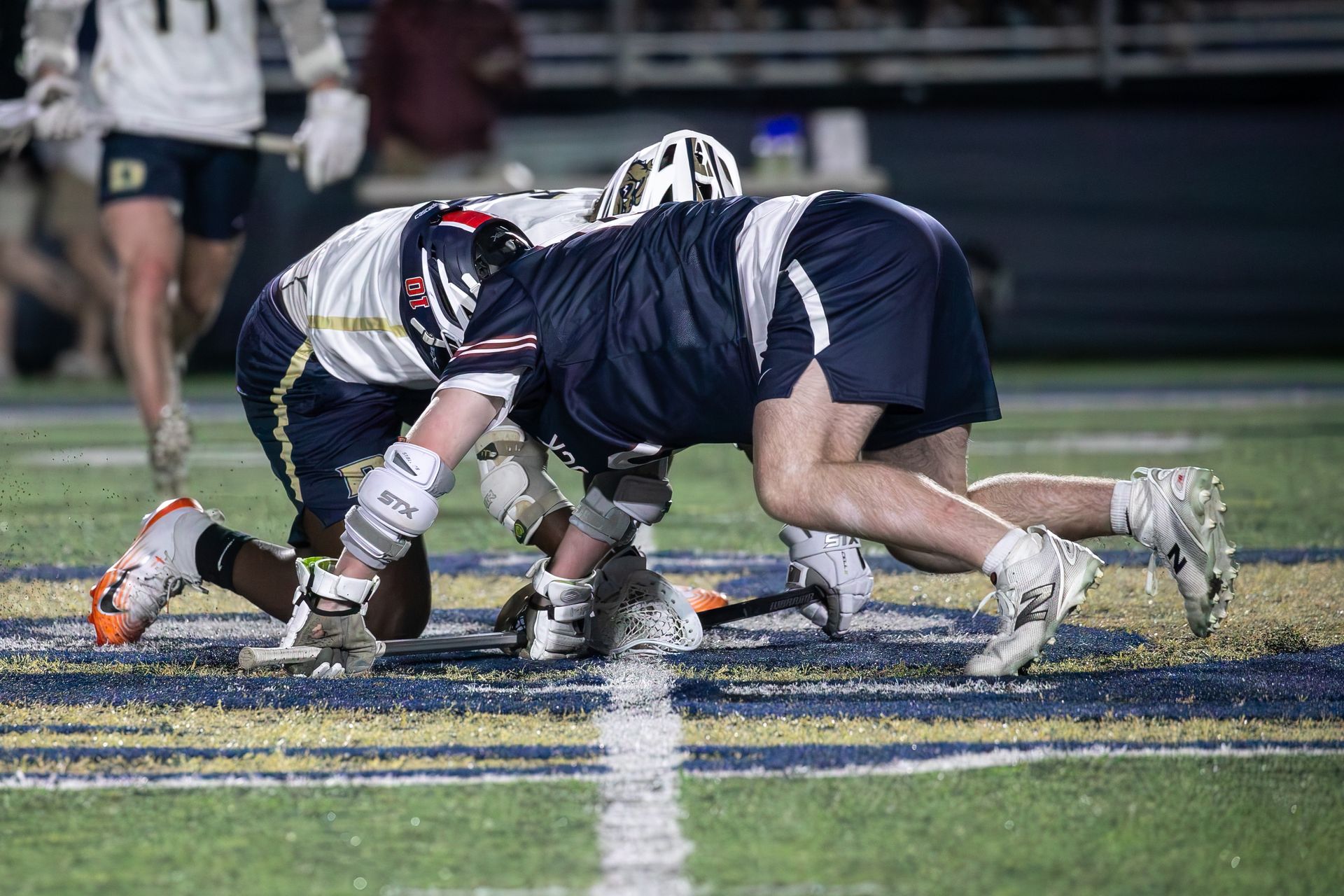 Two lacrosse players in navy and white helmets clash low on a field at night.