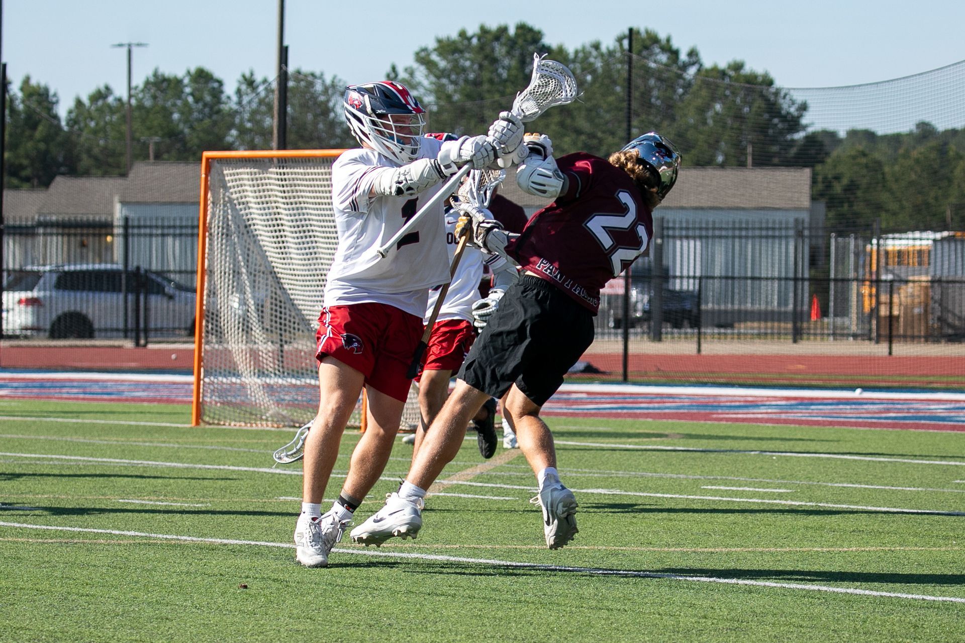 Two lacrosse players collide near the goal, one in white and red, one in black and red.