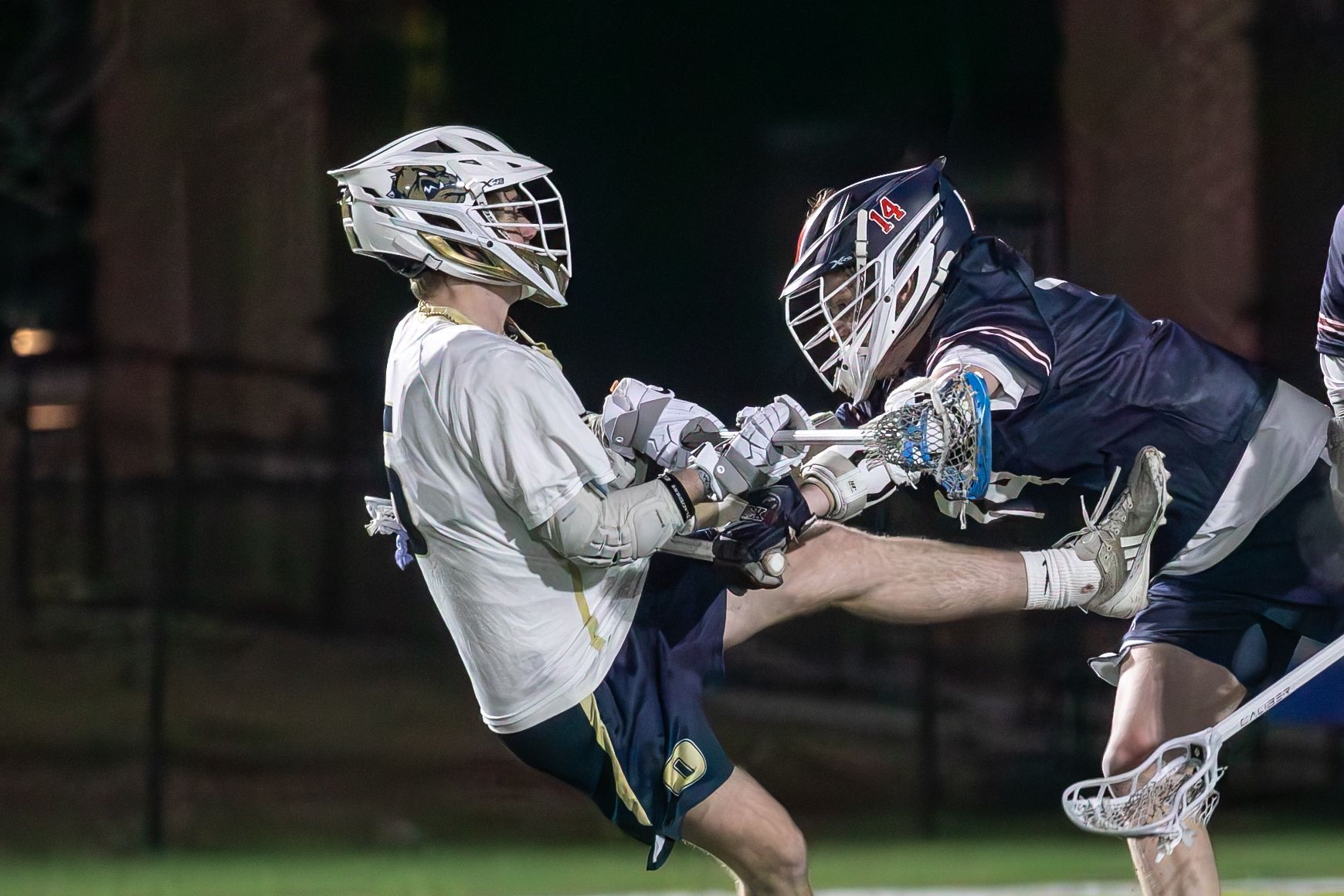 Two lacrosse players collide during a nighttime game, one in white and one in black.