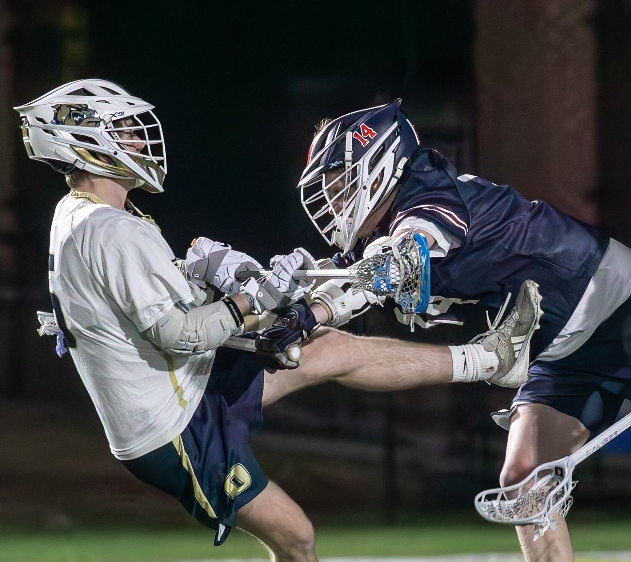 Two lacrosse players in navy and white clash sticks midair during a night game.