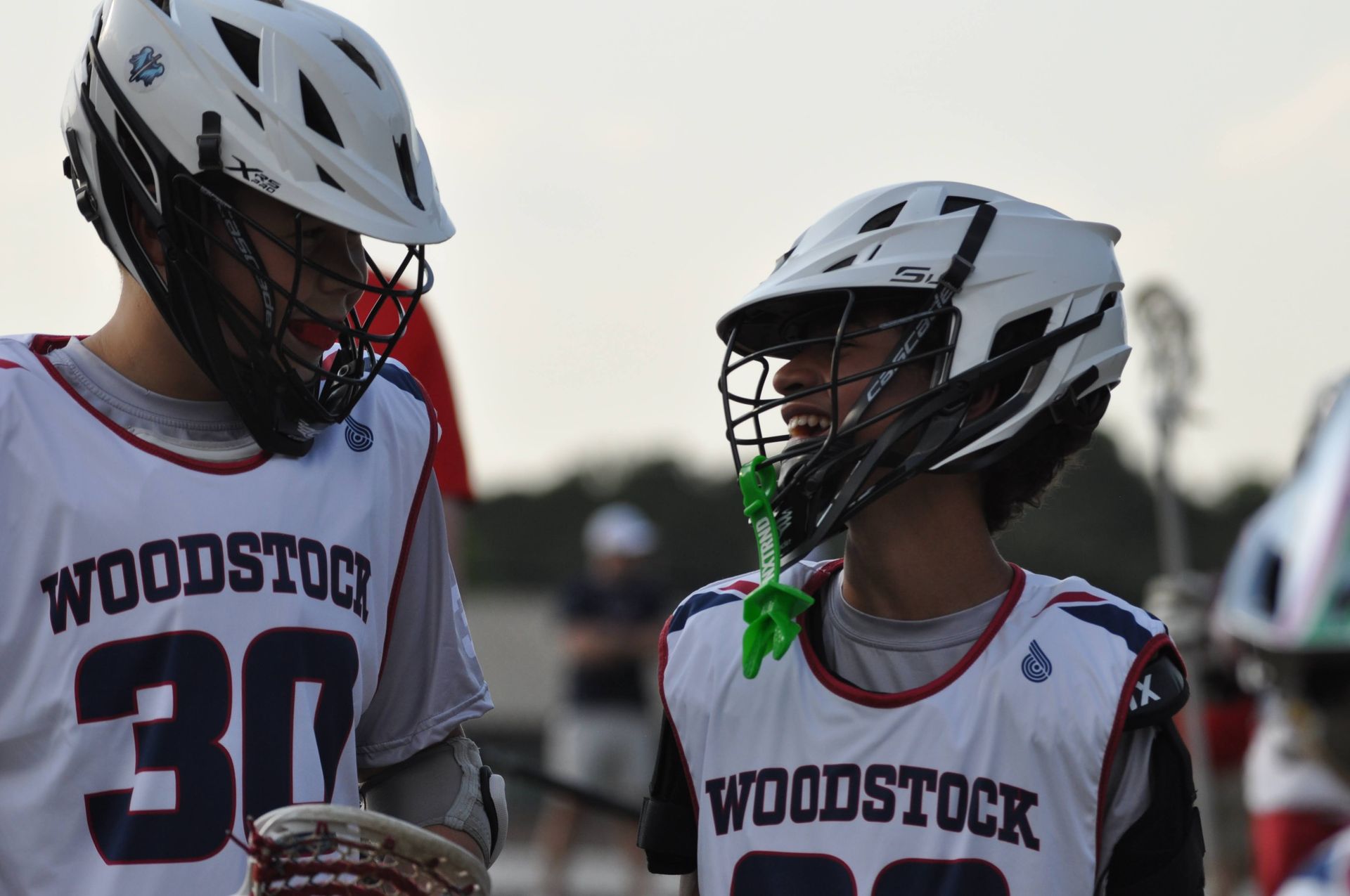 Two Woodstock lacrosse players in white helmets and jerseys talk on the field at dusk