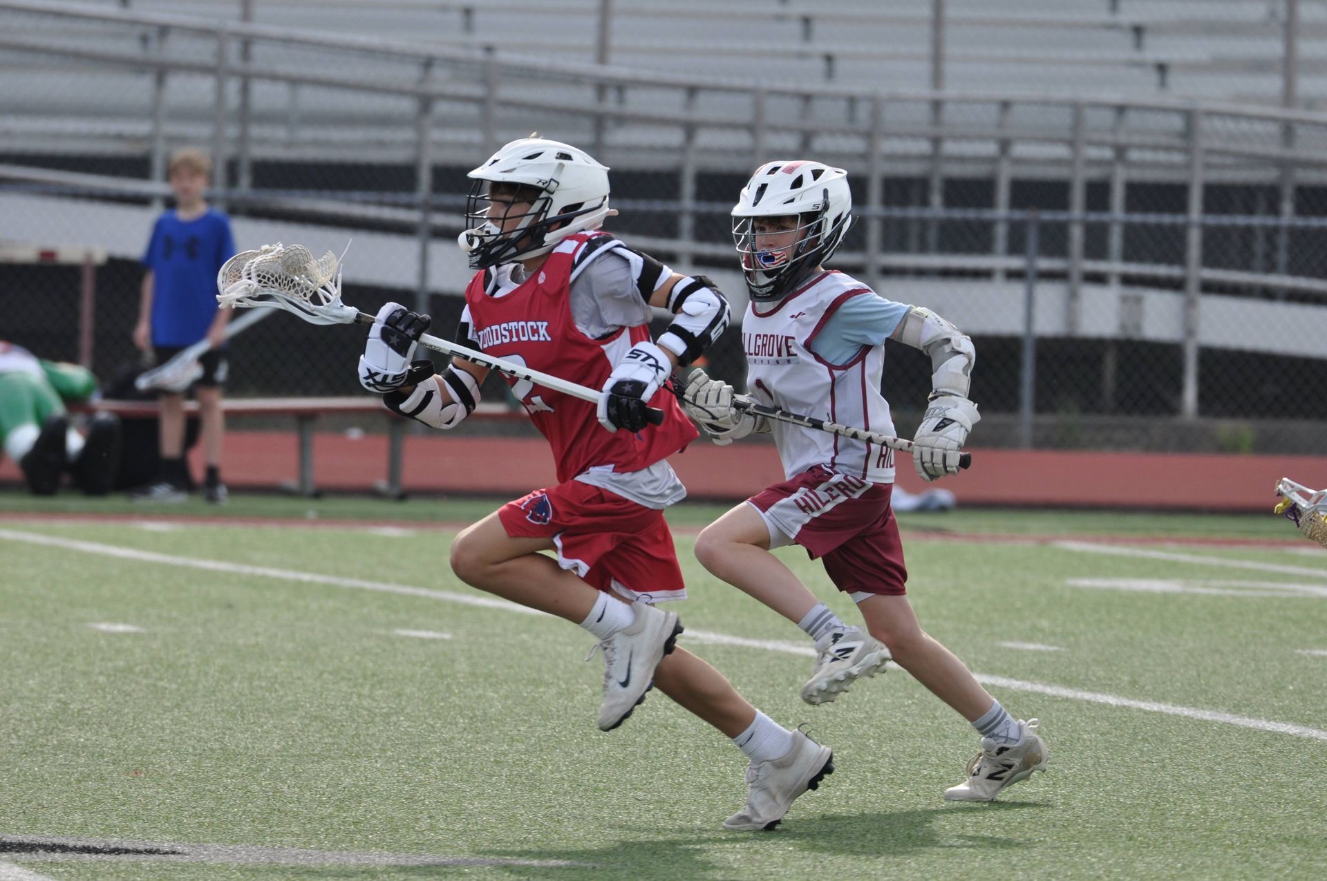 Two lacrosse players sprint on a field, one in red leading with the ball while a white-clad defender chases.