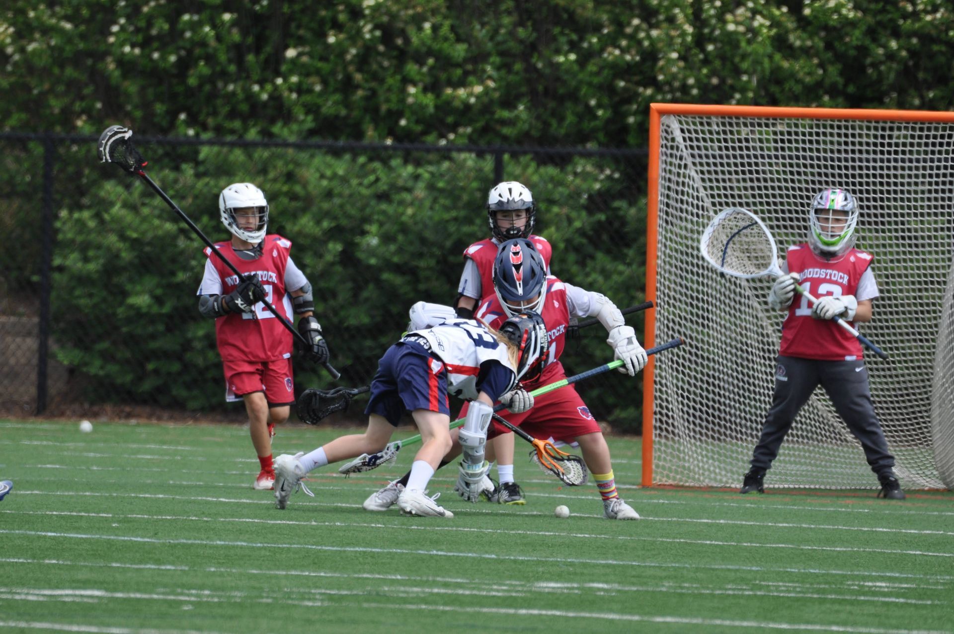 Lacrosse players battle near the goal, with a goalie in red defending on a green field.