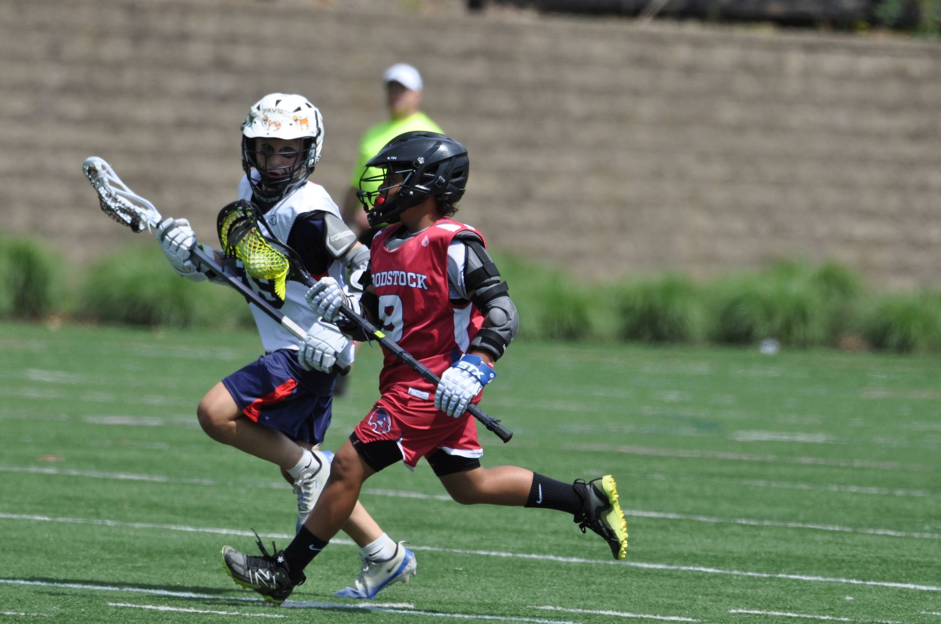 Two lacrosse players sprint on a field, one in red and one in white, both chasing the ball.