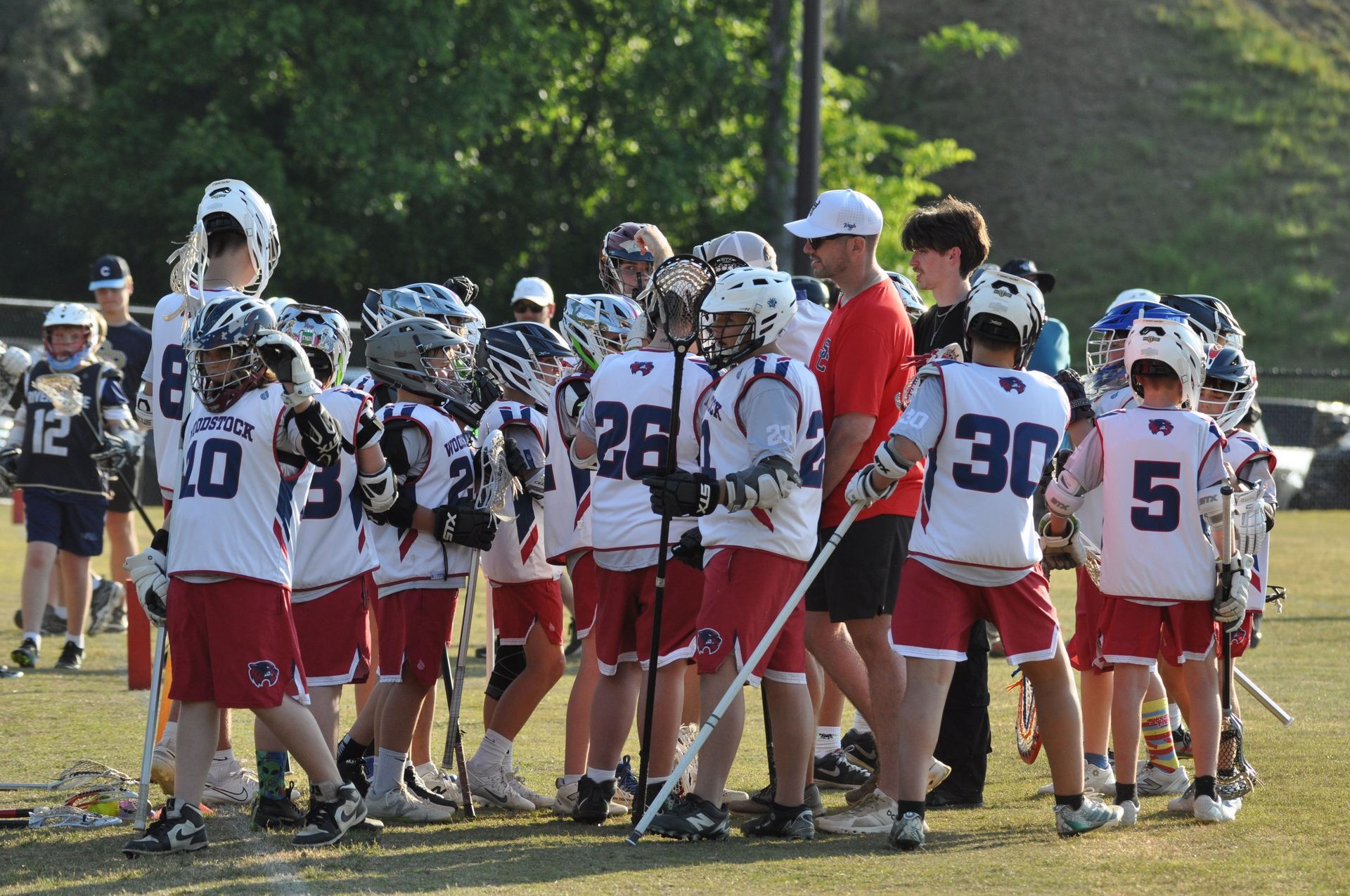 Youth lacrosse team huddles on a sunny field, wearing white jerseys and red shorts.