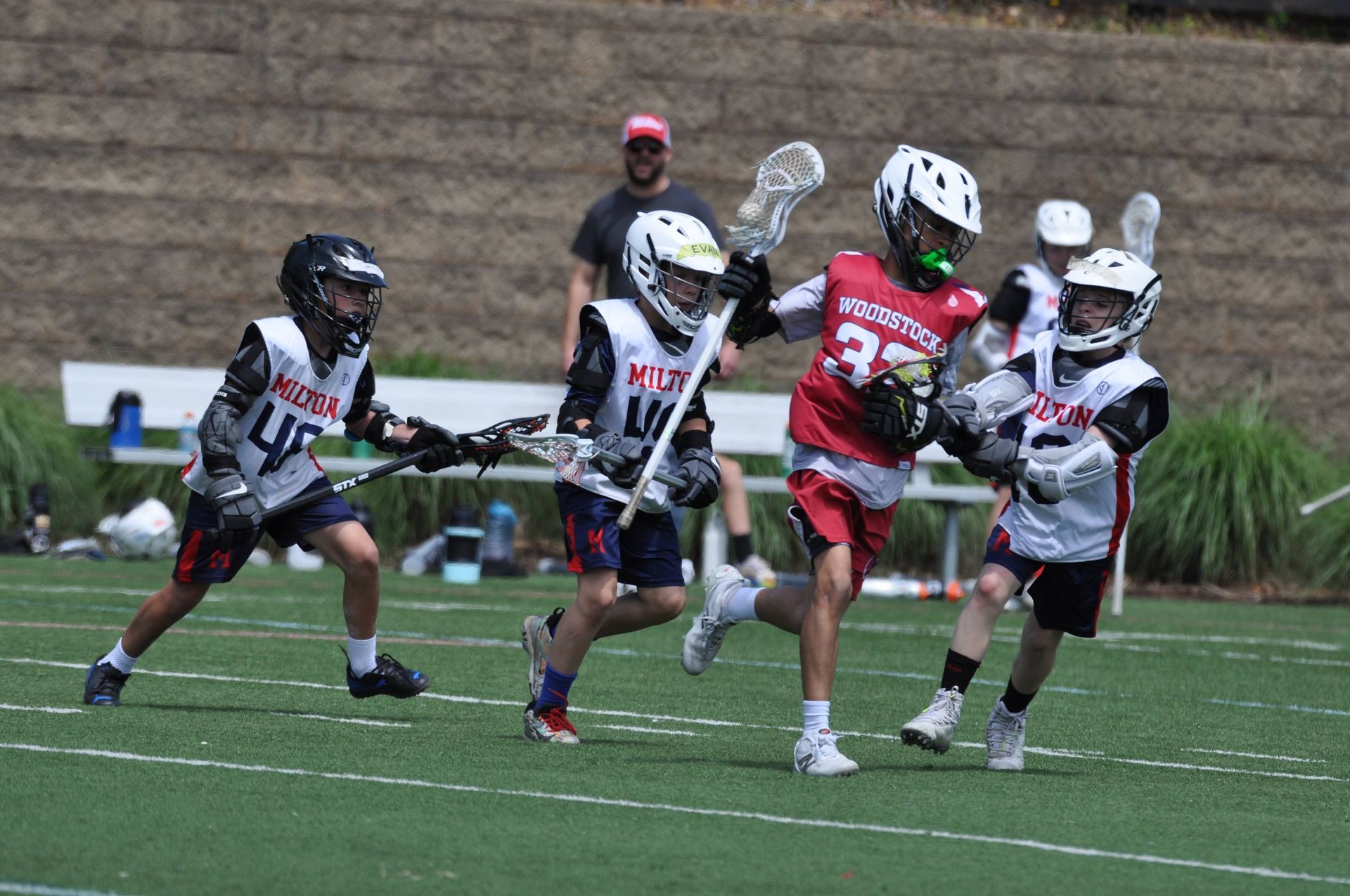 Youth lacrosse players sprint across a field during a game, with a coach watching in the background.