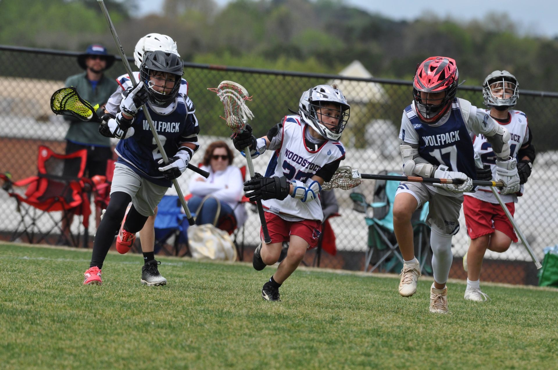 Youth lacrosse players sprint across a grassy field during a game, with spectators watching behind them.