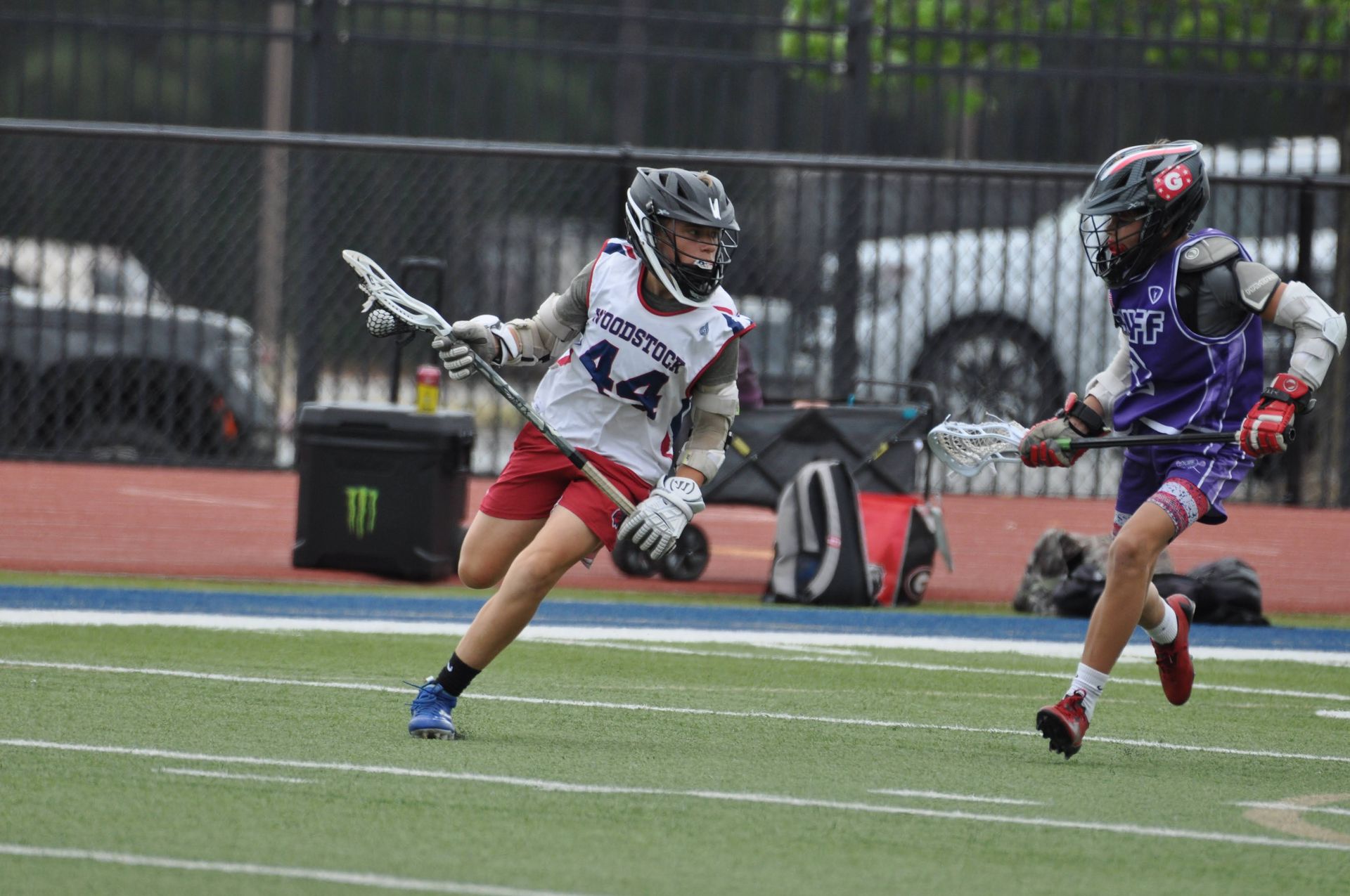 Two lacrosse players in helmets and pads chase the ball on a wet field during a game.