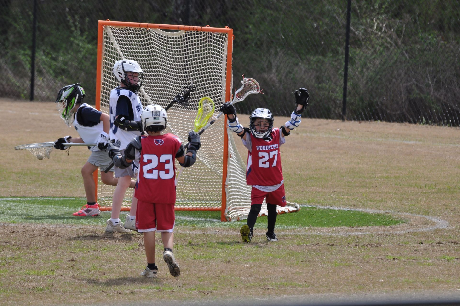 Youth lacrosse players in red celebrate near the goal on a field