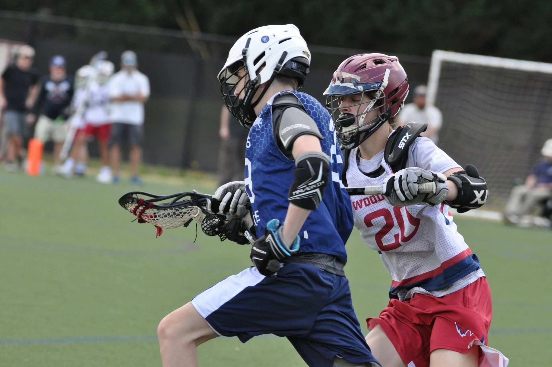 Two lacrosse players in blue and white sprint on a field, one in red and white closely defending.
