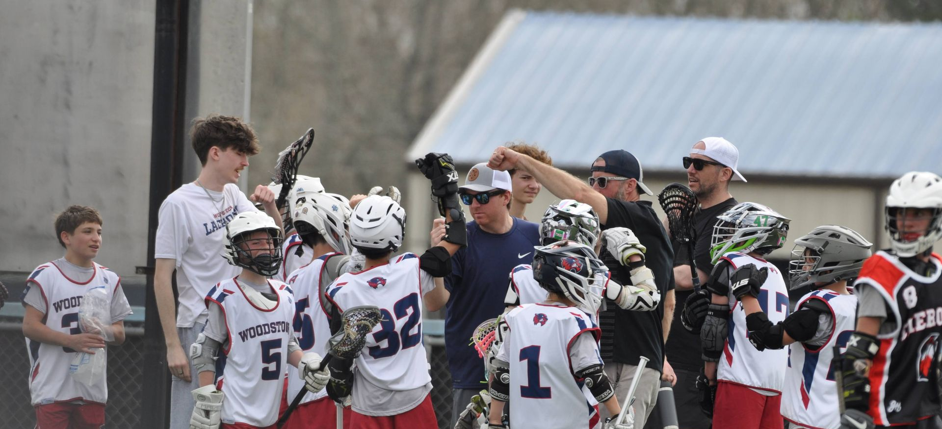 Youth lacrosse players in helmets celebrate with raised sticks on a field after a game.