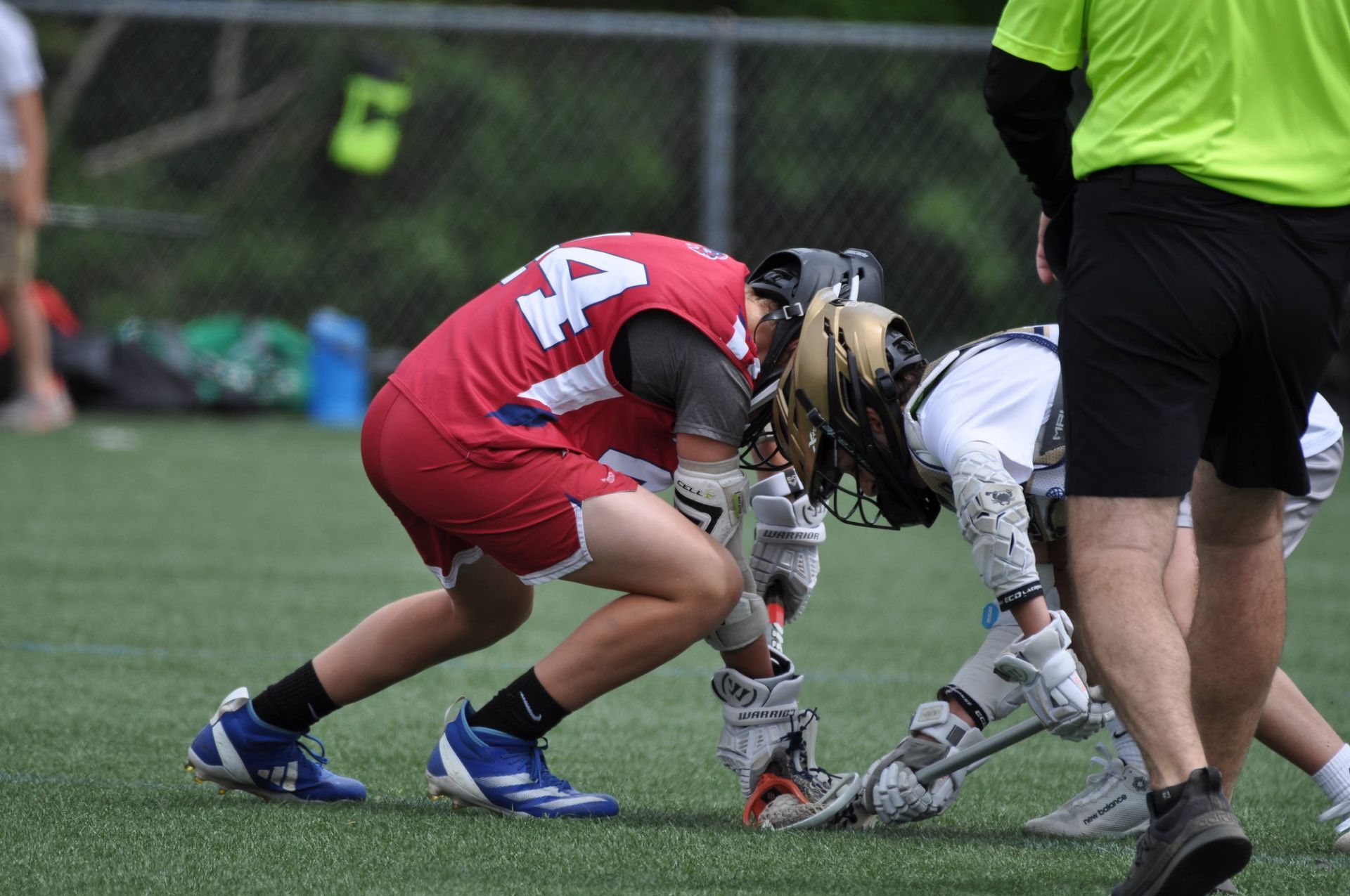 Two lacrosse players crouch faceoff on a turf field, one in red and one in white, with a referee nearby.