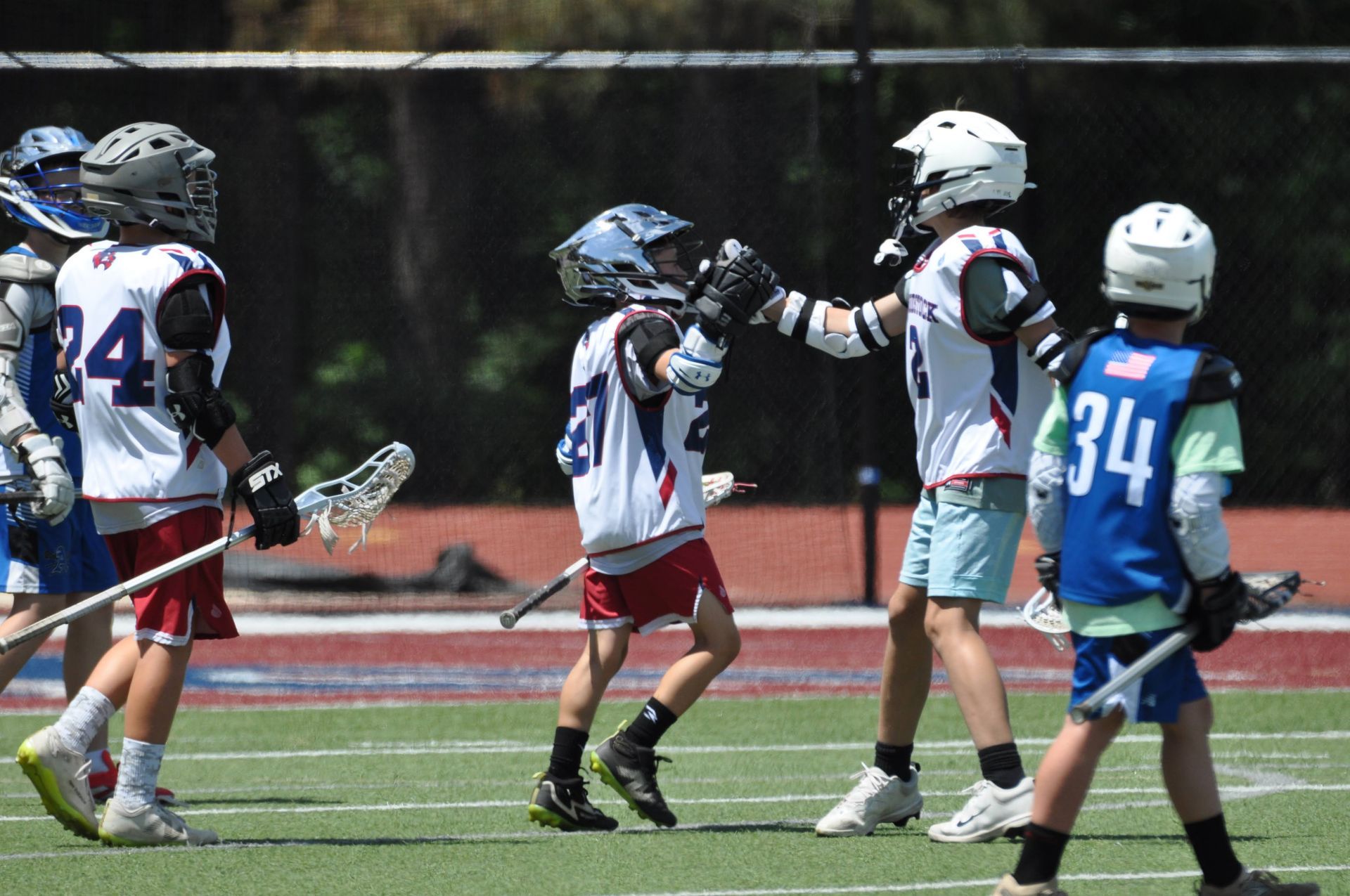 Youth lacrosse players in helmets gather on a field, with one player celebrating at center.