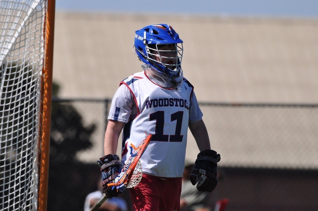 Lacrosse player in blue helmet and white jersey stands near the goal on a field.