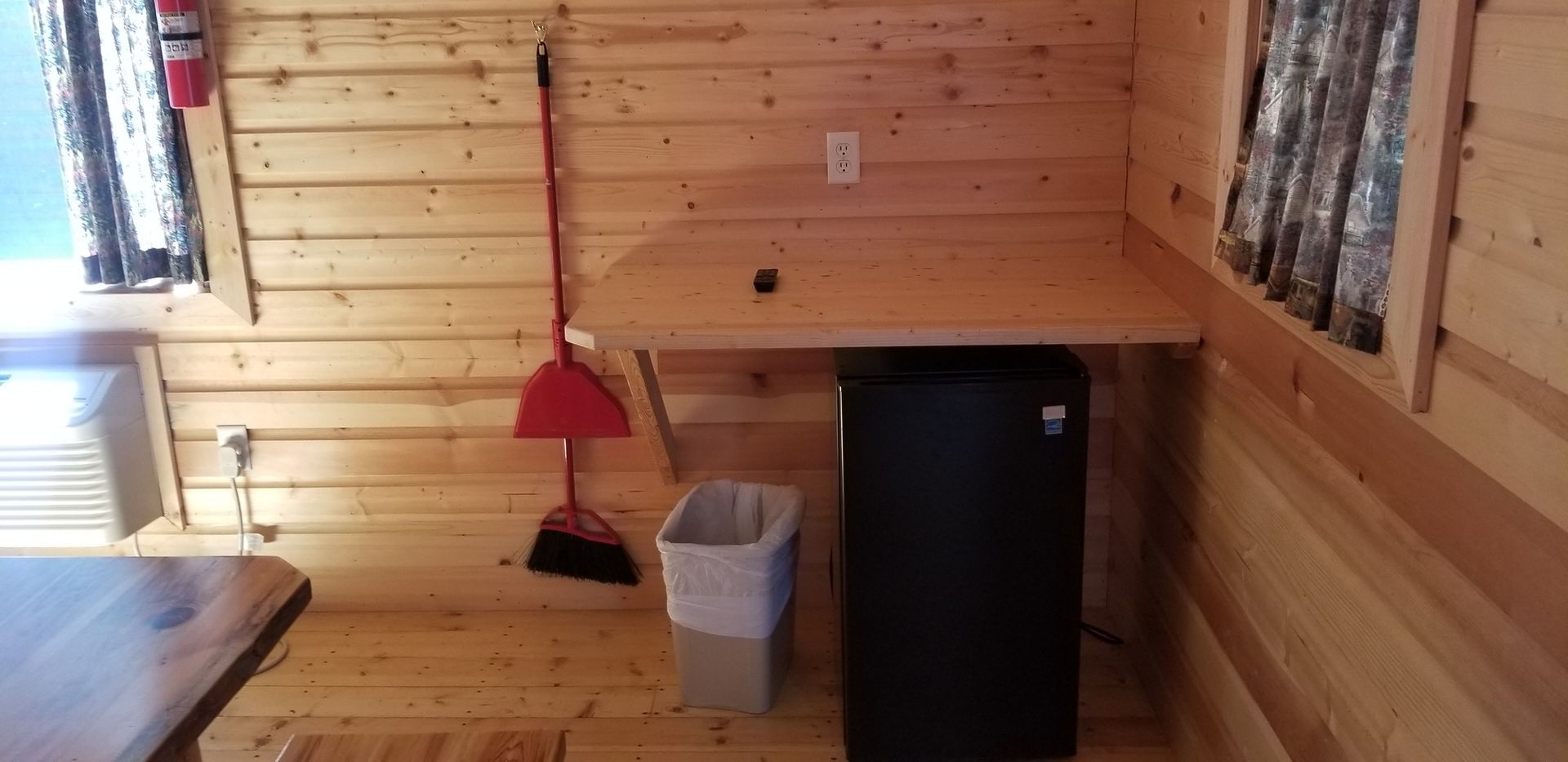 A small refrigerator is sitting under a wooden table in a wooden cabin.