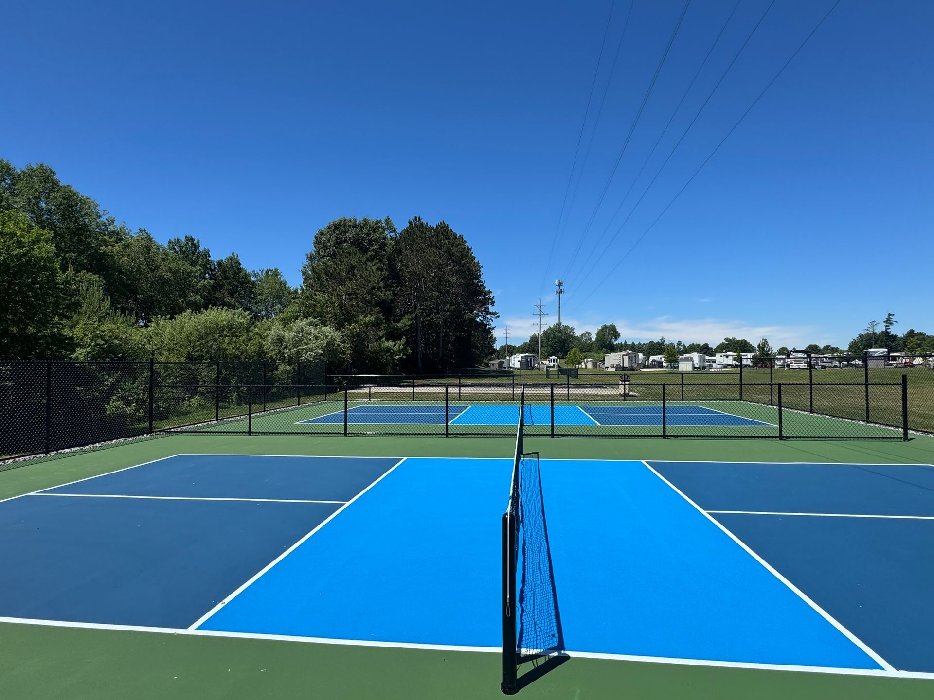 Two blue and green pickleball courts with black nets and fencing, with RVs parked beyond under a clear blue sky.