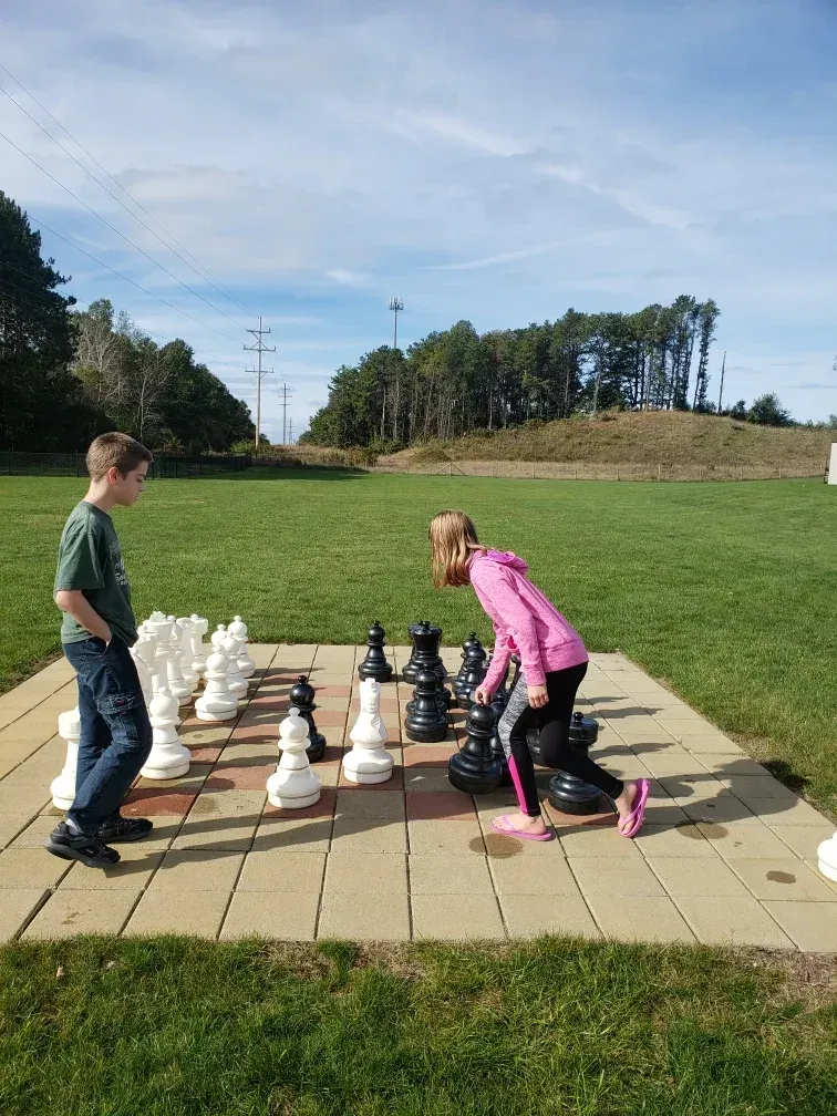 A boy and a girl are playing giant chess in a park.