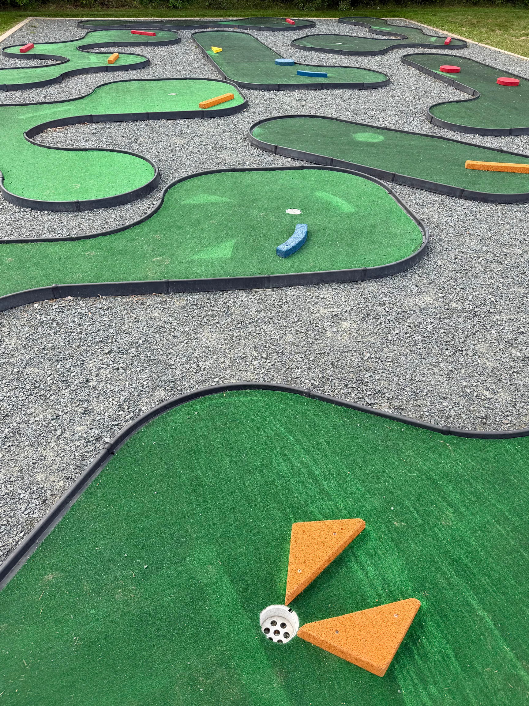 Mini golf course with winding green turf holes, colorful foam obstacles, and gravel walkways between greens.
