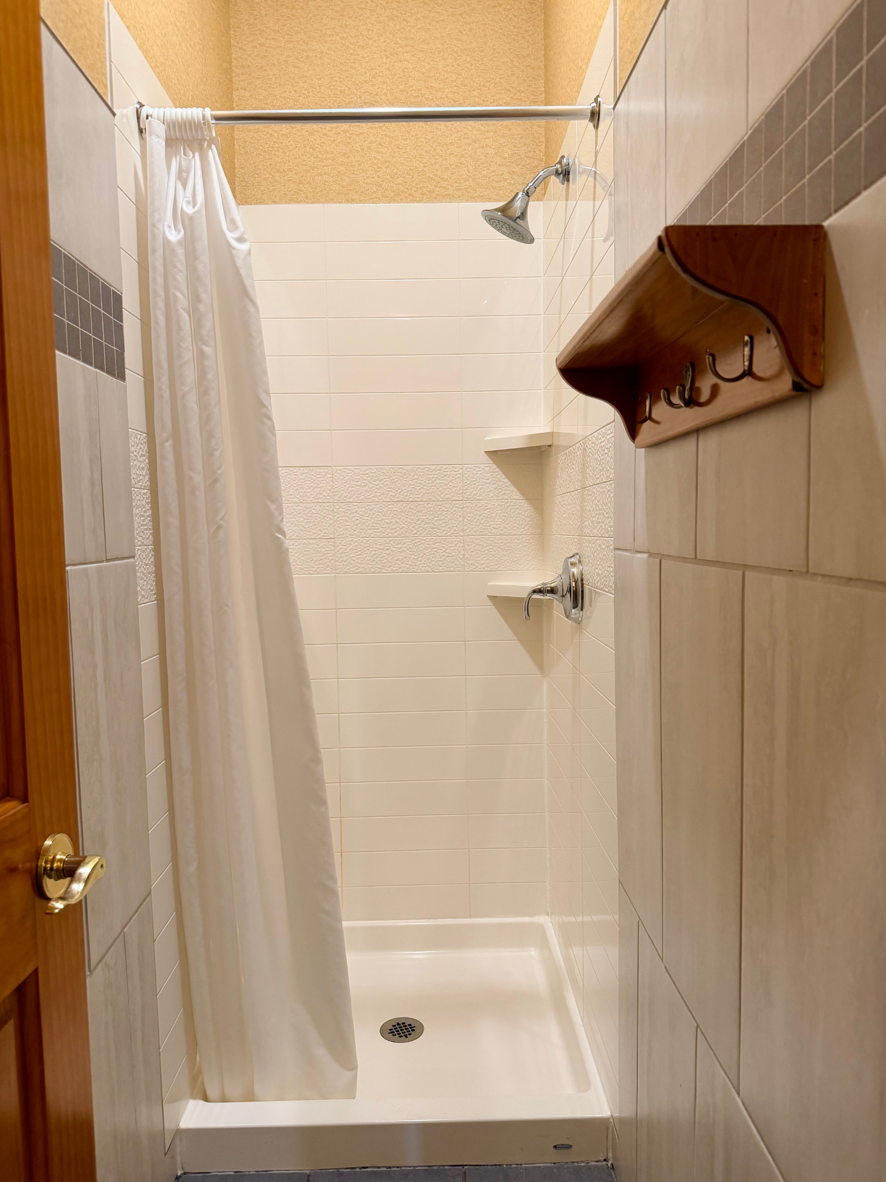Clean private shower stall with tile walls, chrome fixtures, corner shelves, and a wooden door outside