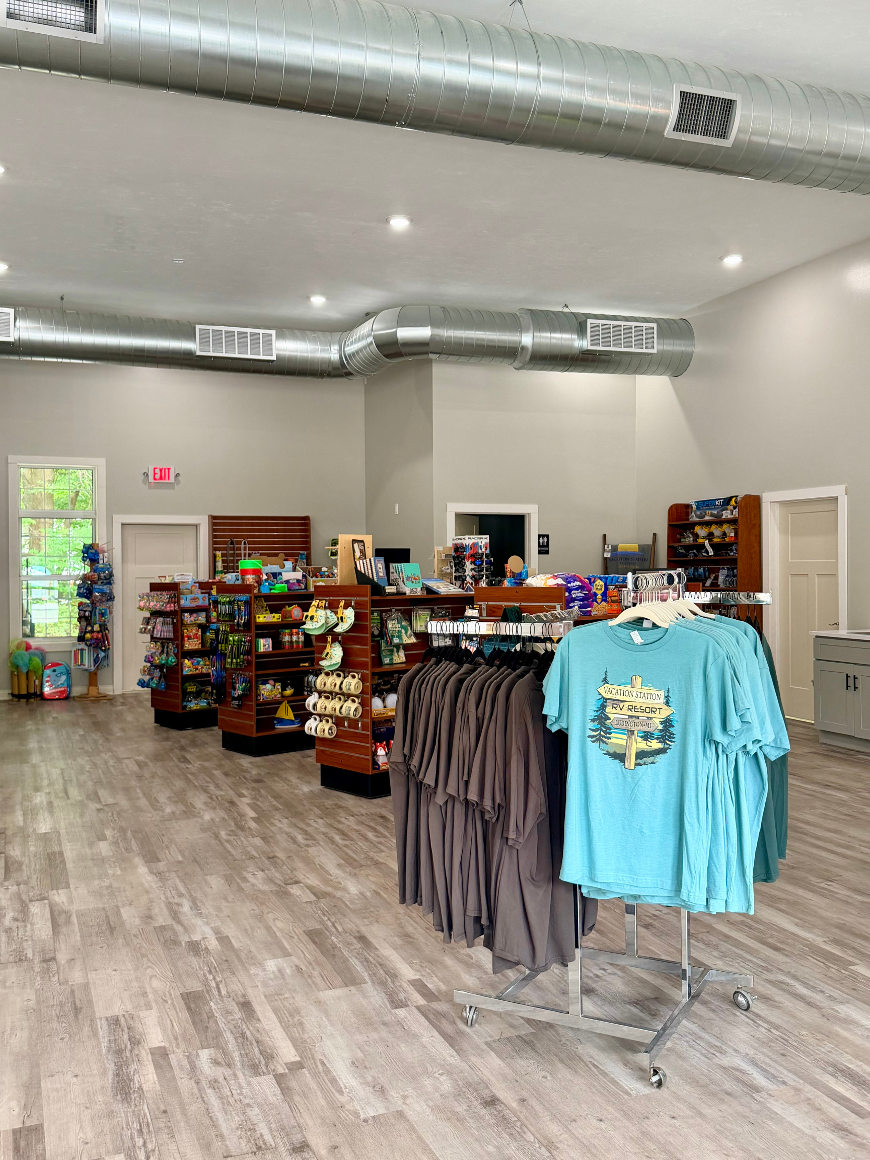 Camp store interior with shelves of snacks, toys, and souvenirs, plus a rack of Vacation Station RV Resort t-shirts.