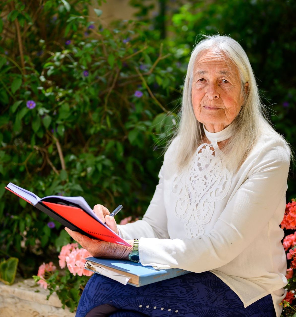 An elderly woman is sitting on a bench holding a book and a pen