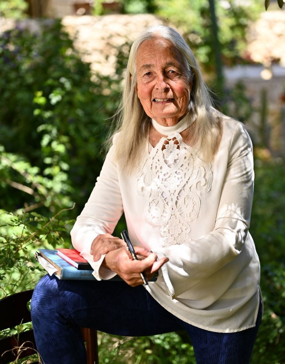 An elderly woman is sitting on a bench holding a pen and a book.