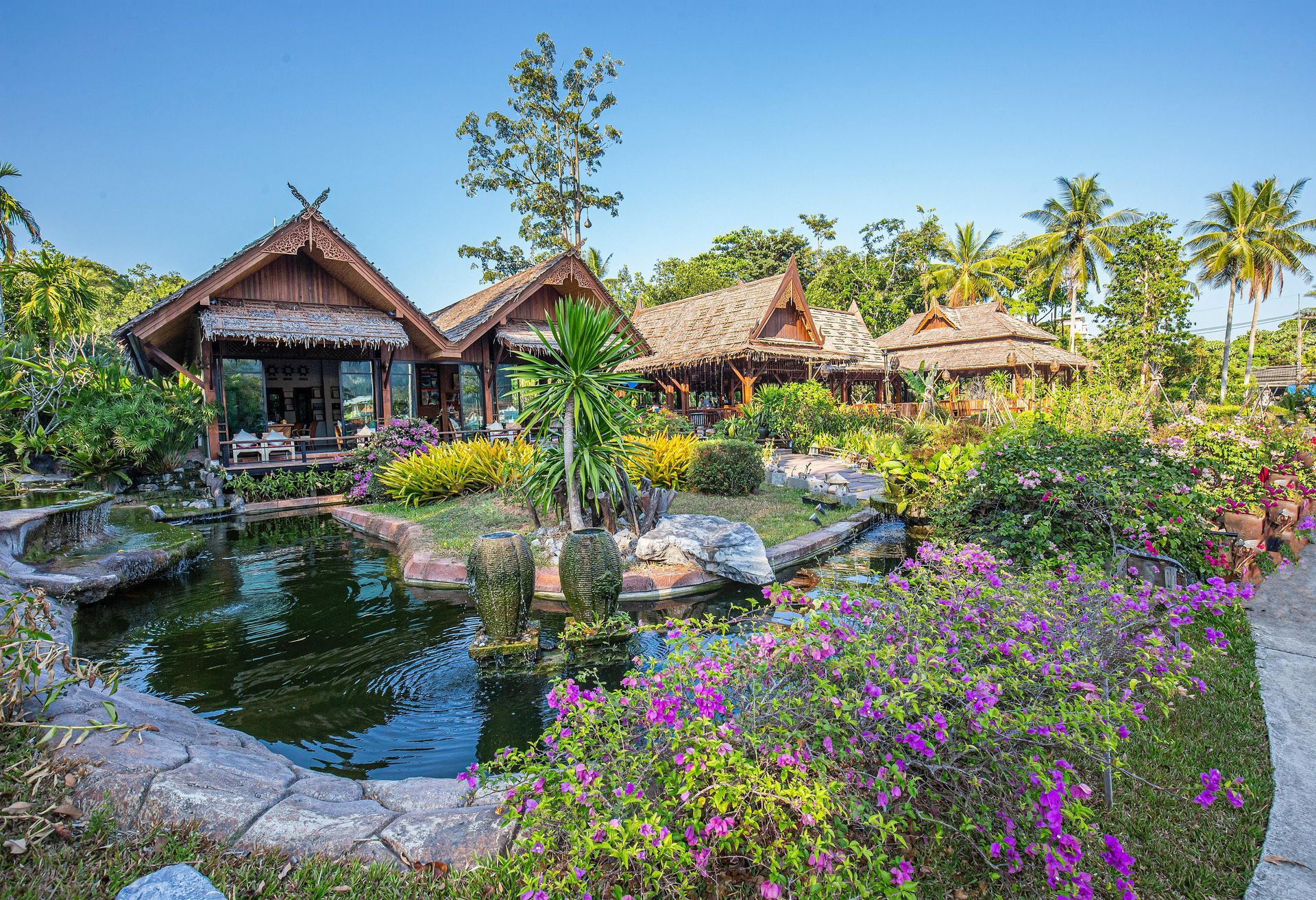 Traditional Thai huts with thatched roofs, surrounded by lush gardens, a pond, and palm trees.