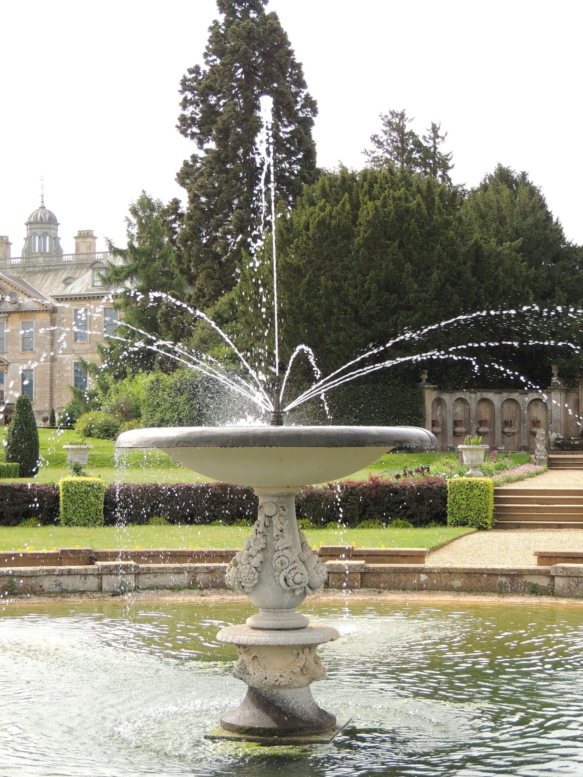 Fountain in a pond, water spraying. Large building and greenery in the background.