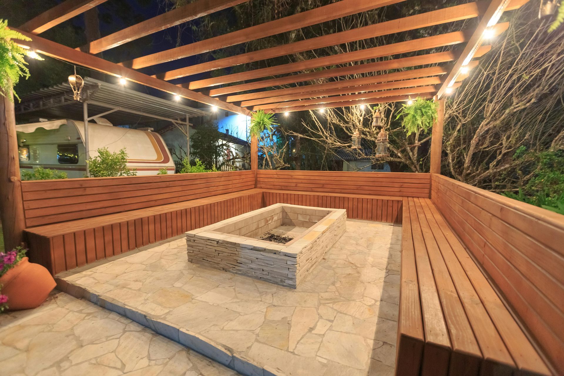 Outdoor seating area with fire pit, wooden benches, and pergola lit by string lights.