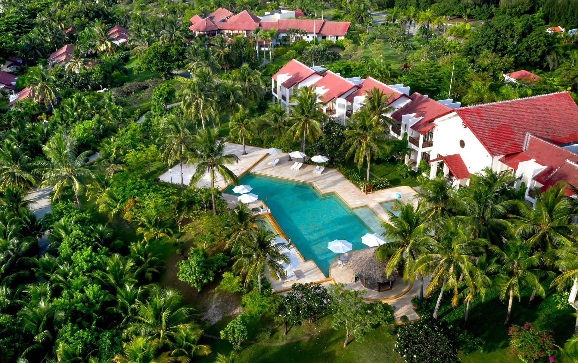 Aerial view of a tropical resort with red-roofed buildings, palm trees, and a turquoise pool