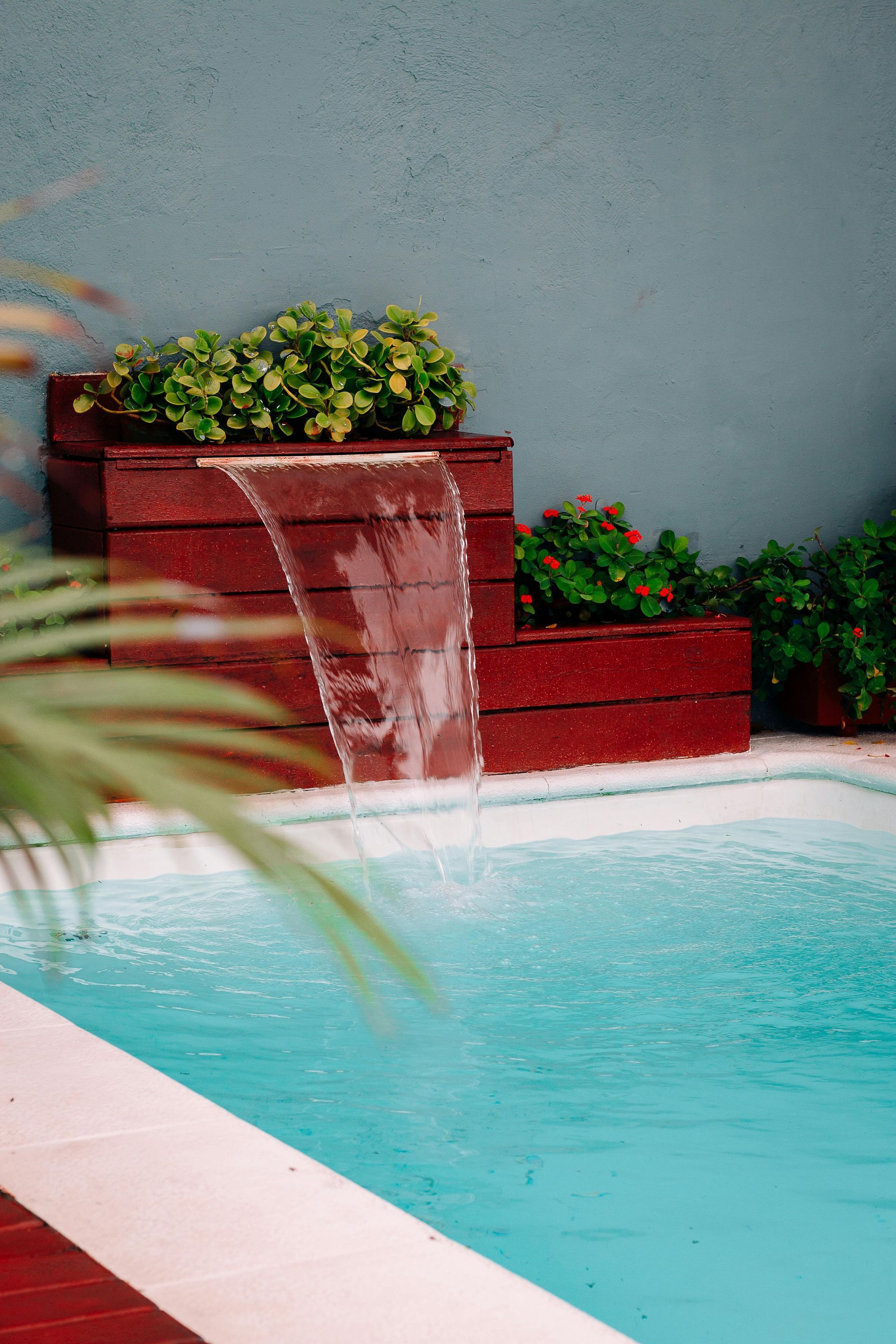 Waterfall feature cascading into a pool, surrounded by plants and red-brown planters against a teal wall.