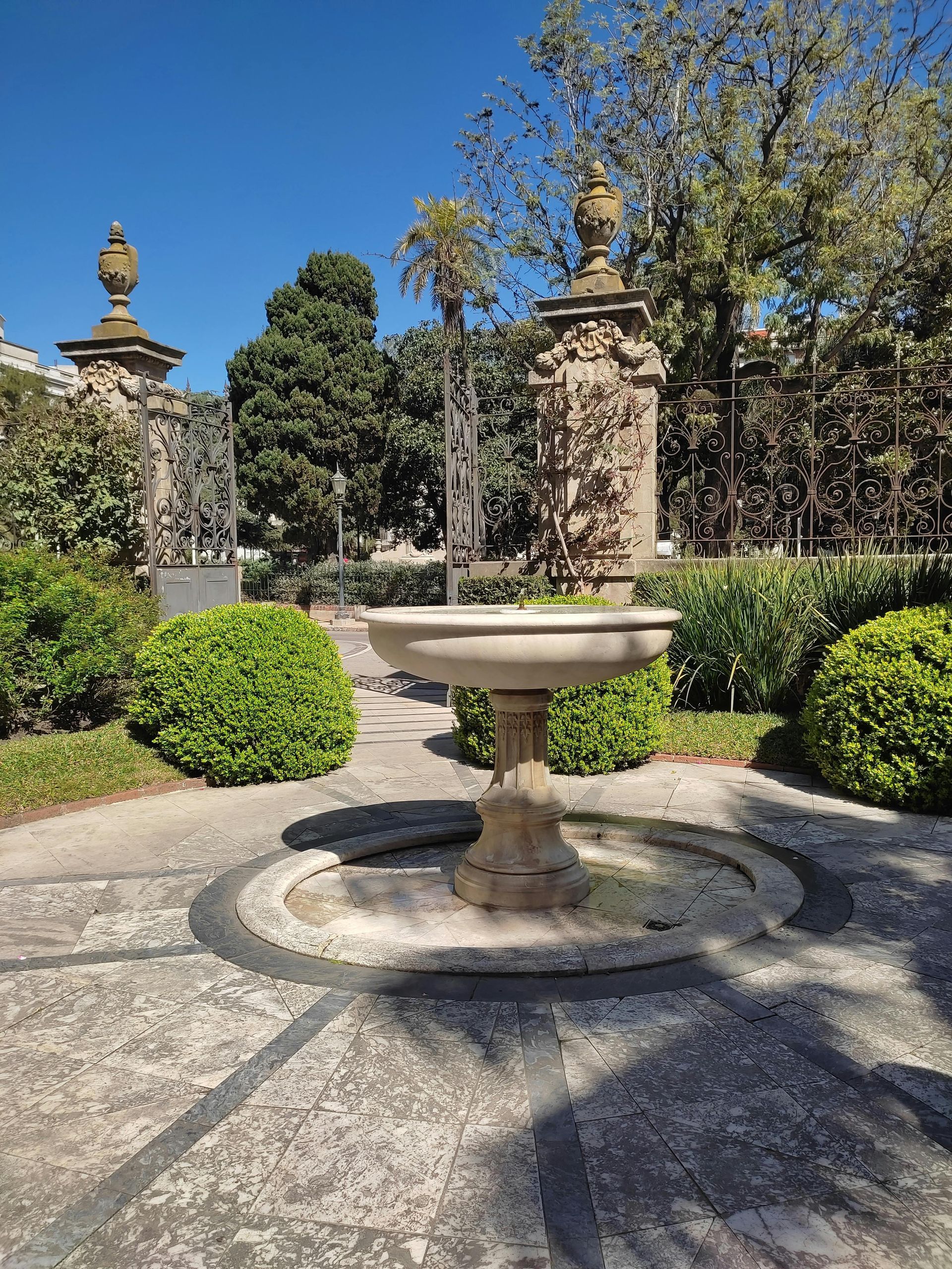 Stone fountain in a garden. Ornamental pillars and greenery surround it, under a bright blue sky.
