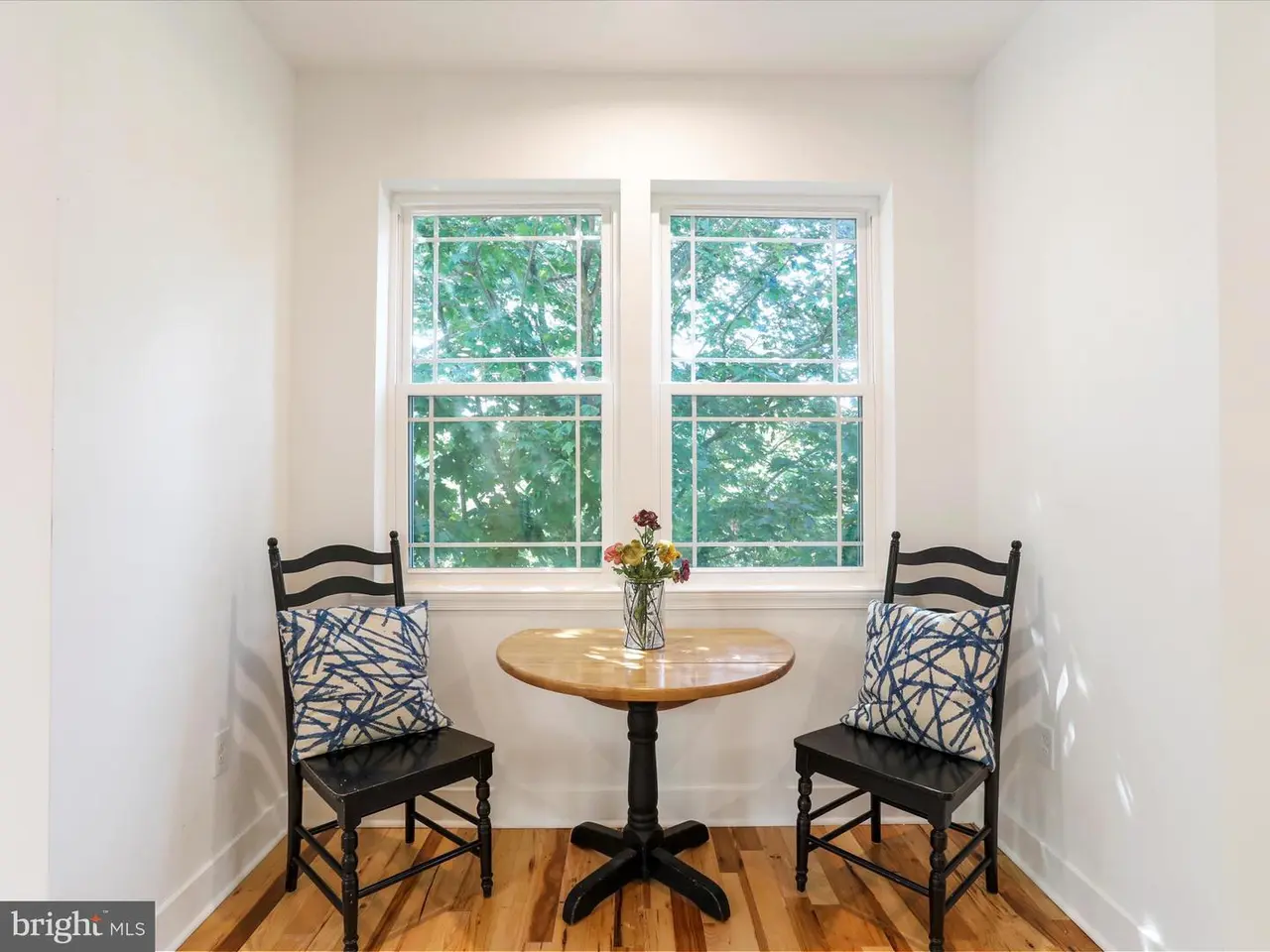 Two black chairs flank a small table in front of a window; flowers in a vase on the table.
