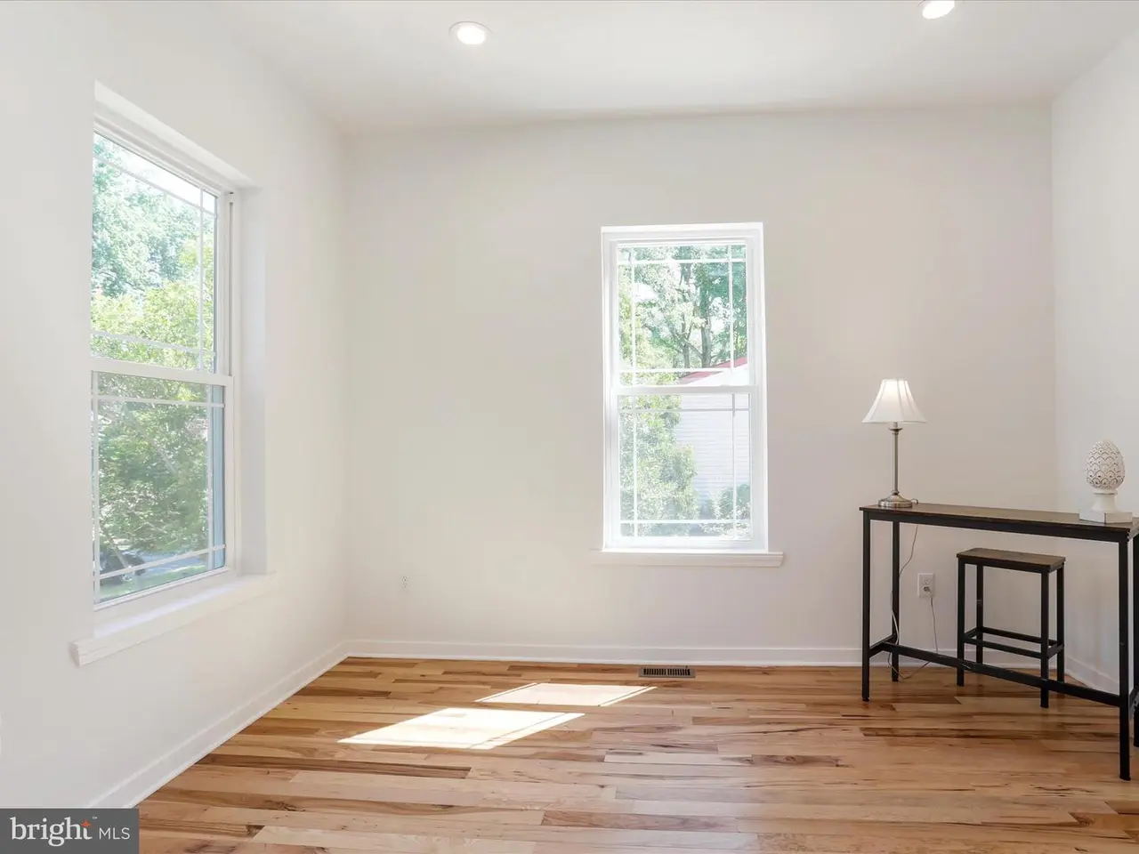 Empty room with hardwood floors, two windows, desk, and lamp. White walls, bright.