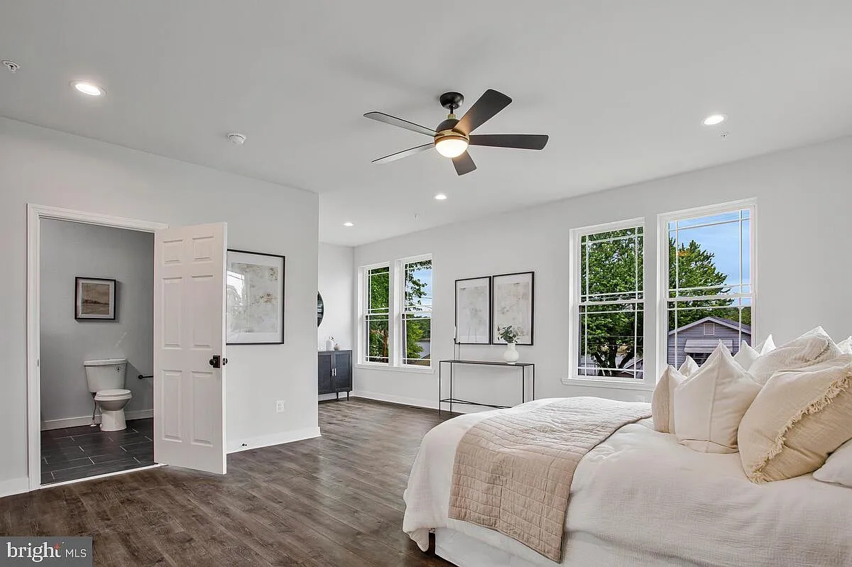 Bright master bedroom with large windows, white walls, and a bed. Adjacent bathroom visible.