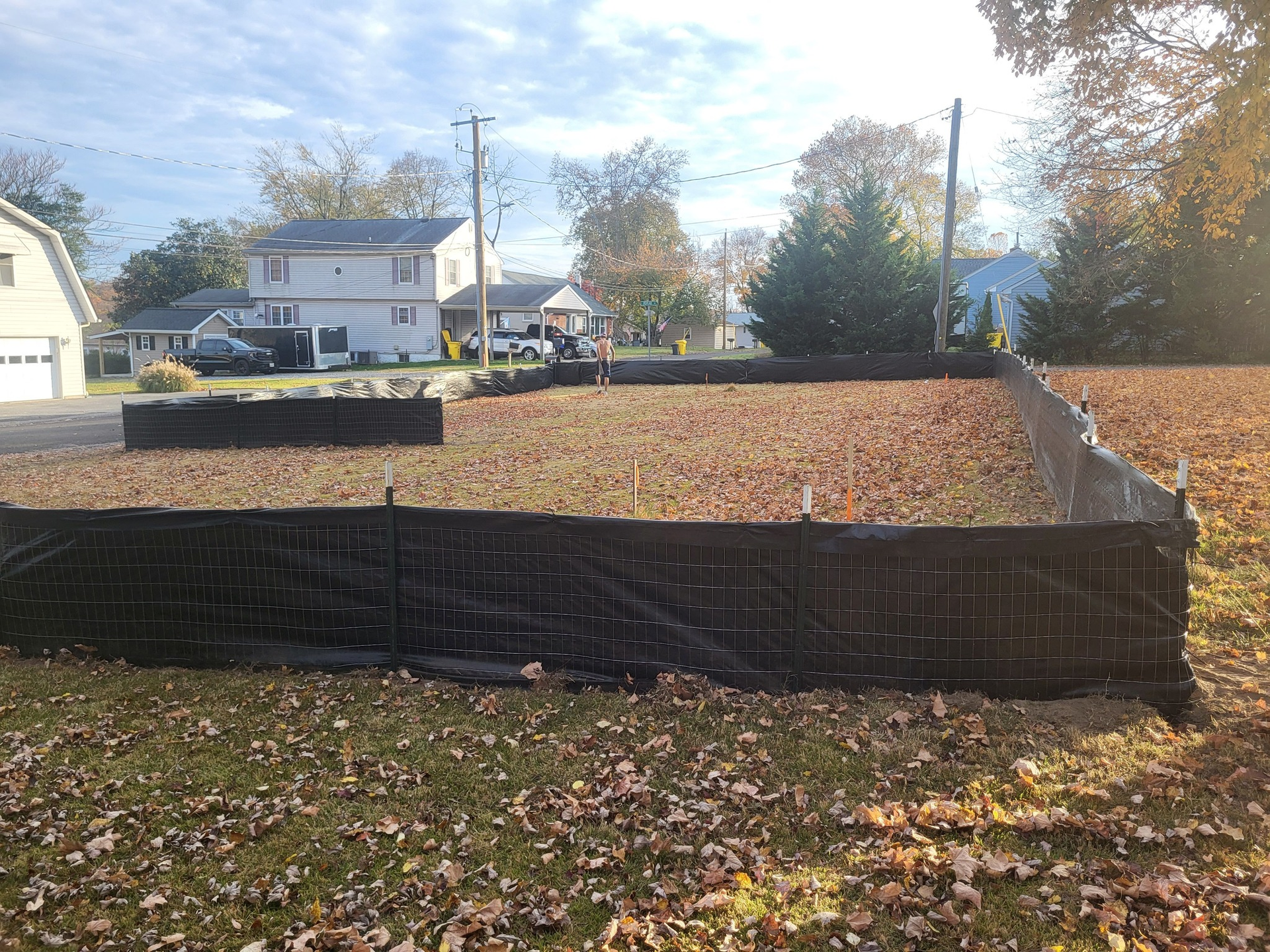 A fenced-in construction site in a residential yard, filled with leaves. Houses and trees in the background.