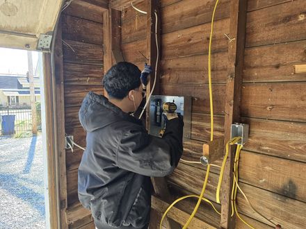 Person working on an electrical panel in a wooden shed, with yellow cables and tools nearby.