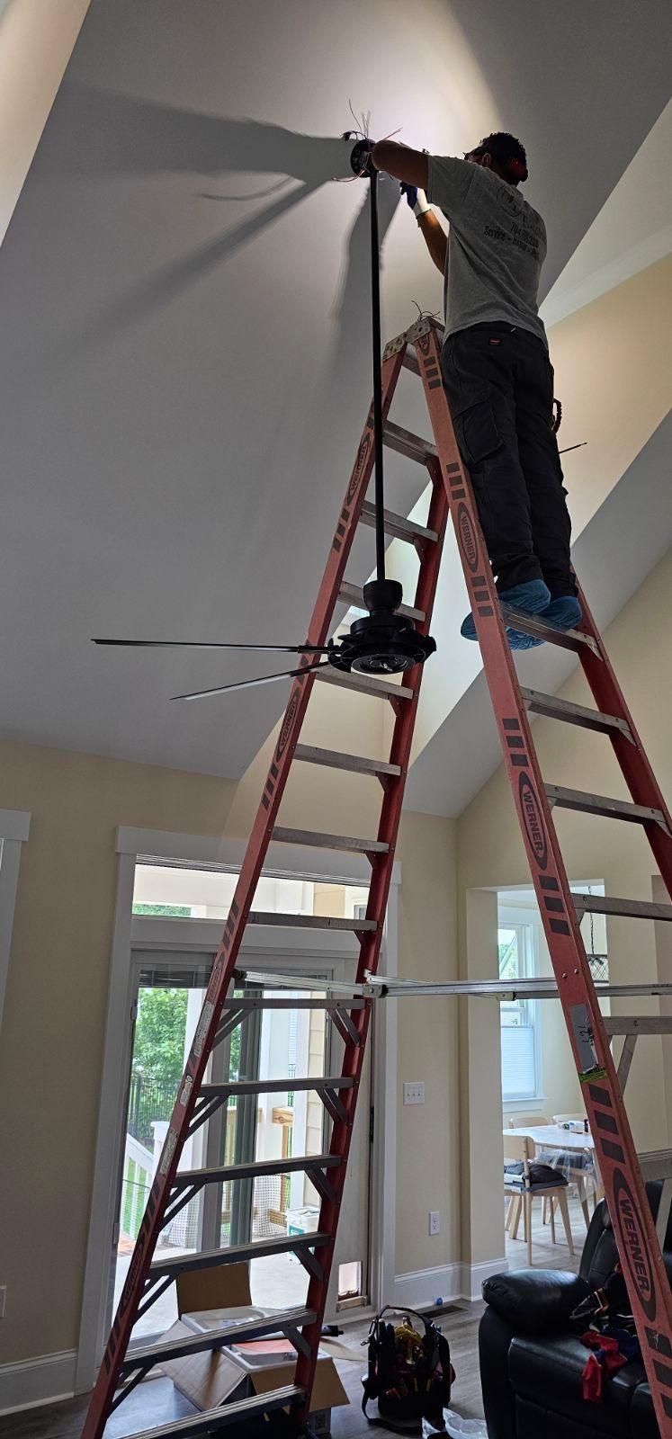 Person on red ladder painting a high white wall near a ceiling fan in a bright room