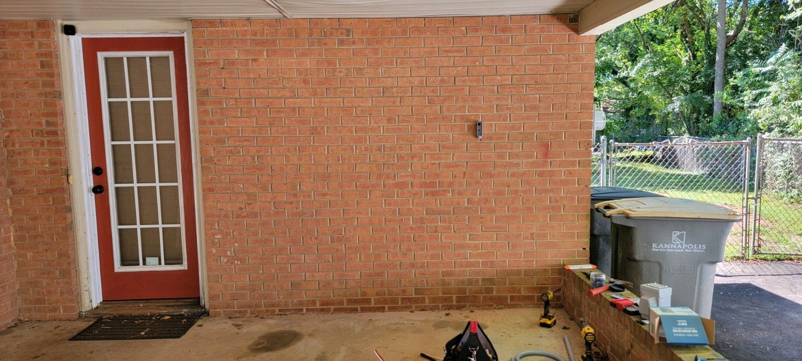 Covered brick patio with a red door, brick wall, and two trash bins on the right.