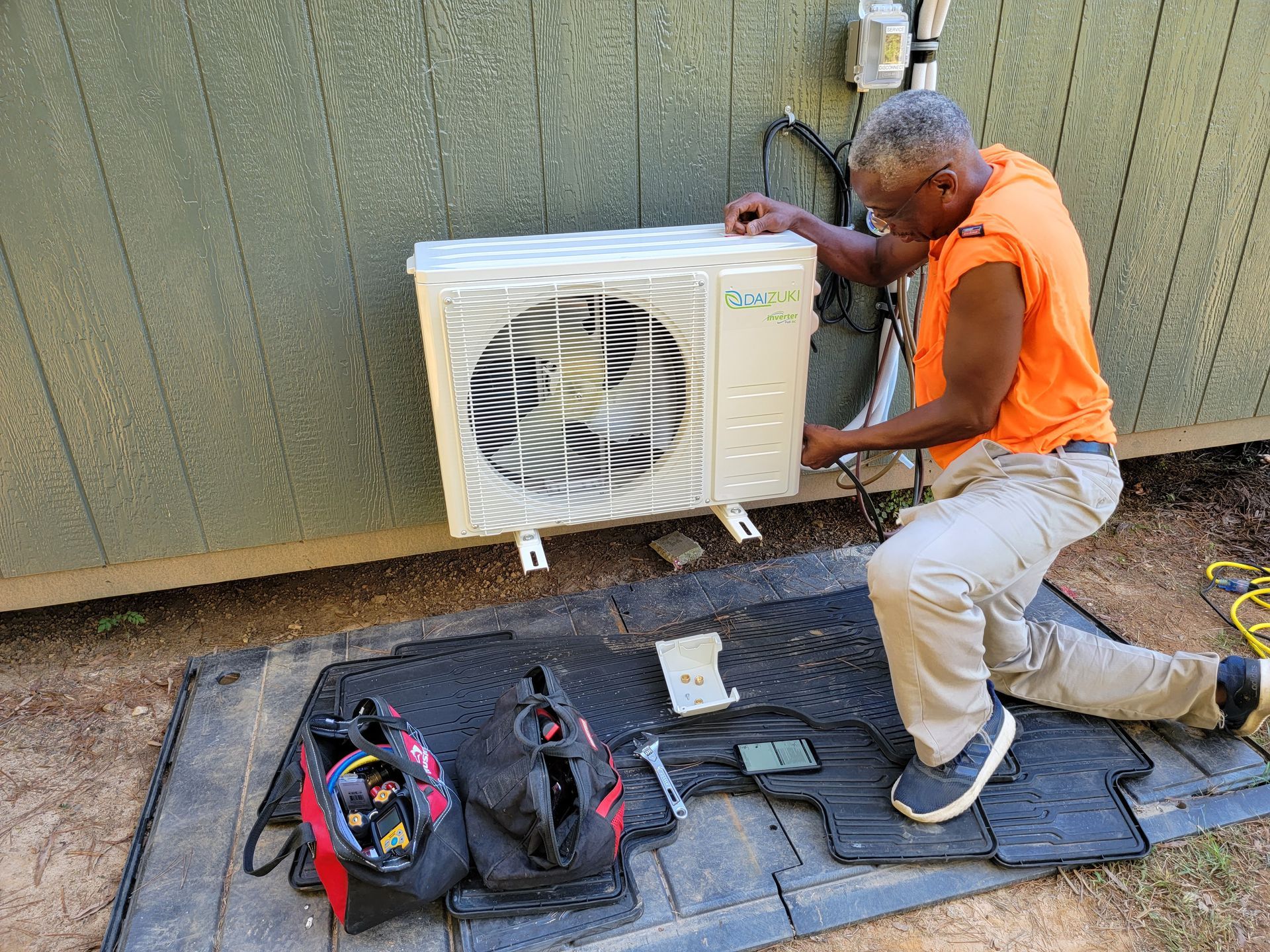 Worker in orange vest kneels by an outdoor AC unit, using tools on a mat against a wall.
