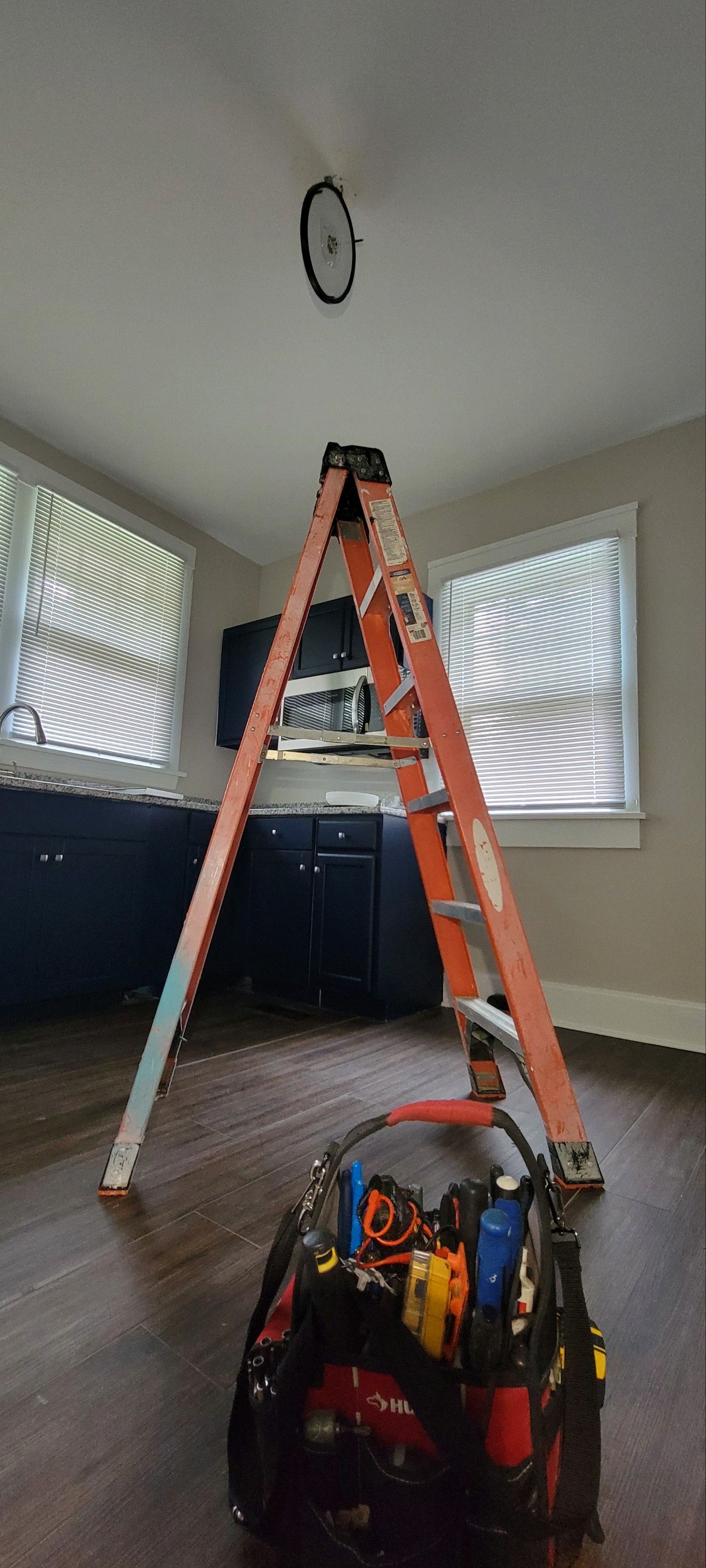 Living room with a tall orange ladder under a ceiling light, and a tool bag on the floor near windows.
