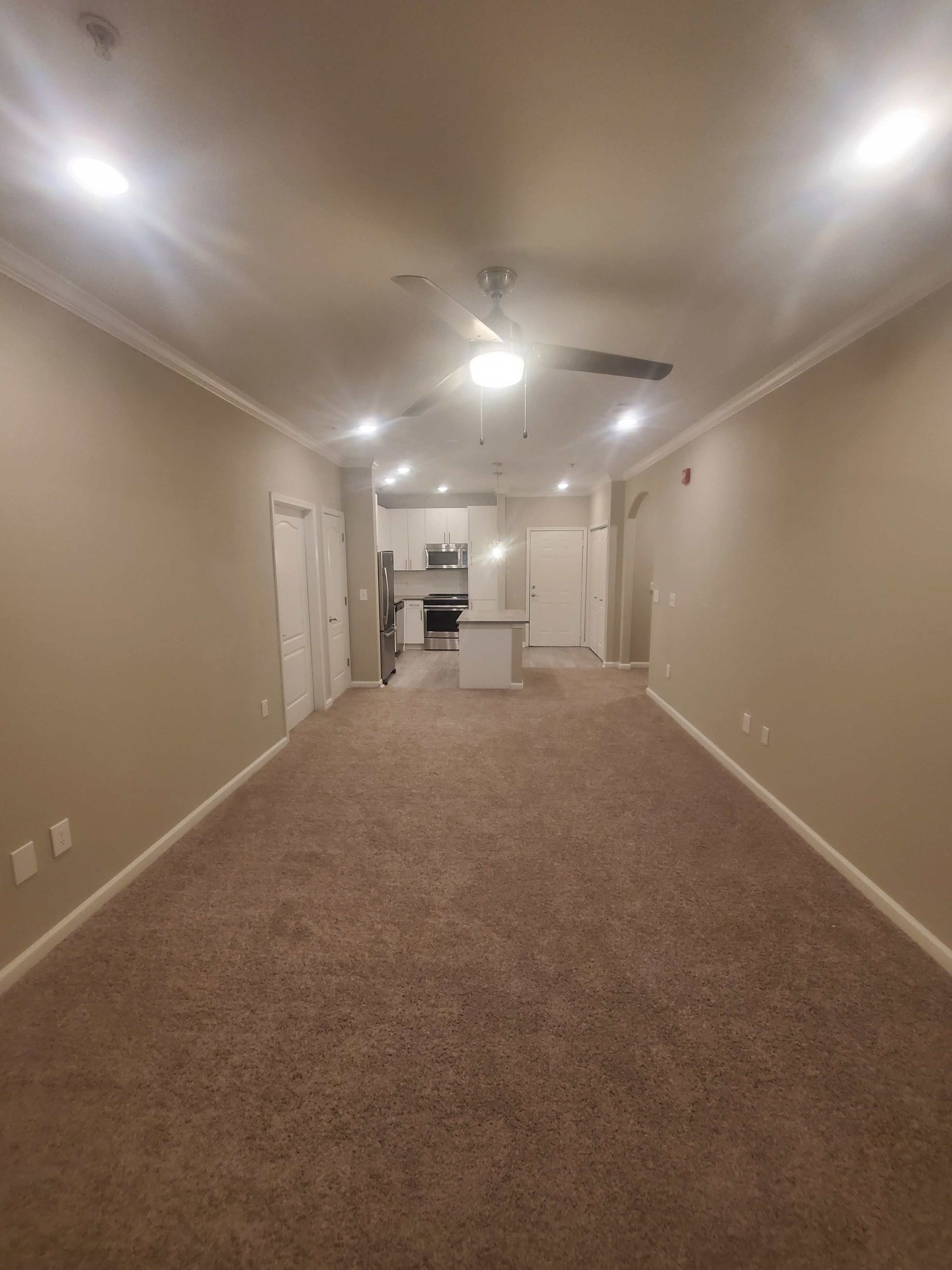 Empty beige carpeted room with recessed lights and a ceiling fan, opening to a kitchen area.
