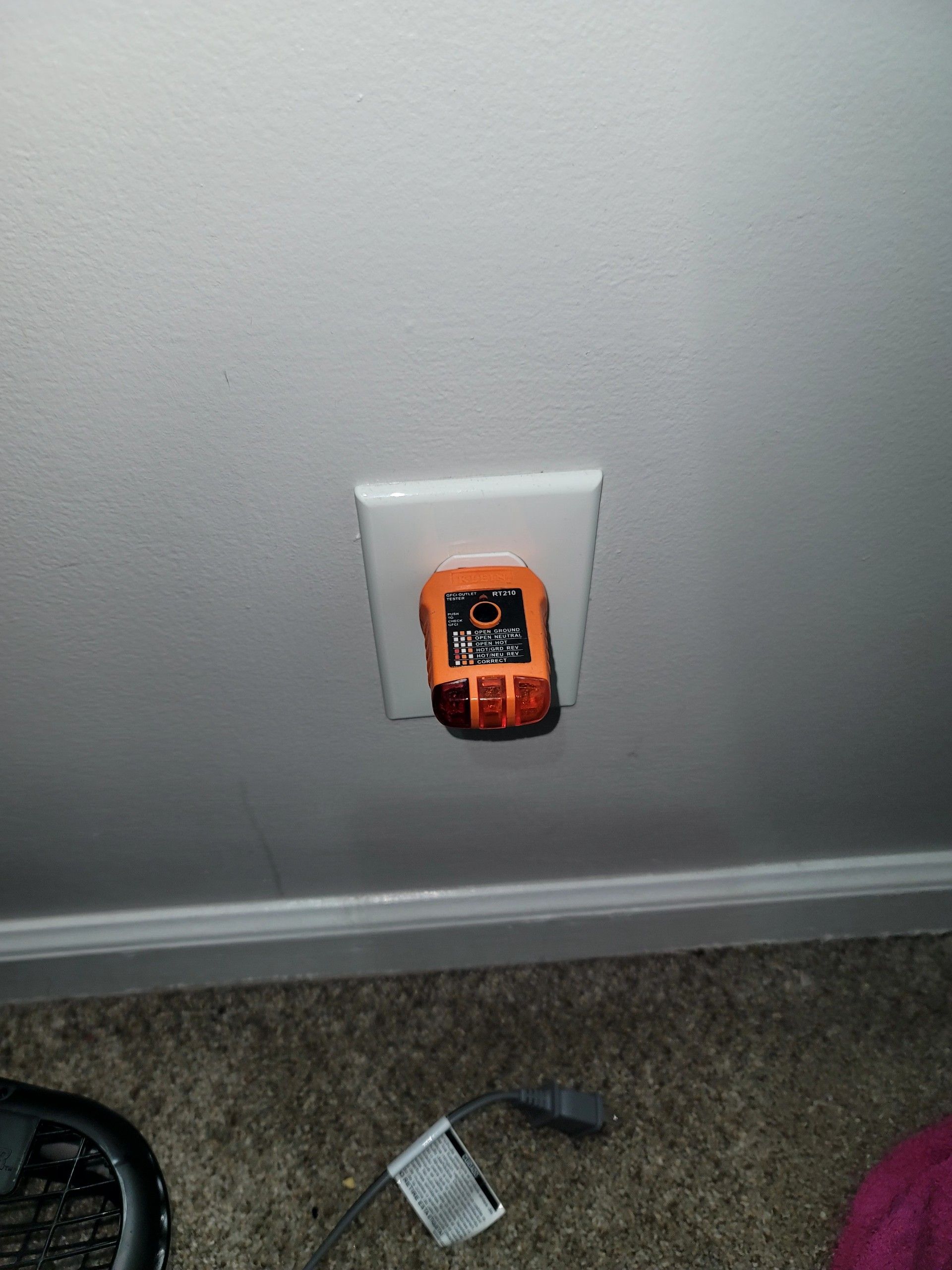 Orange electrical outlet tester plugged into a wall outlet near the floor on carpeted ground.