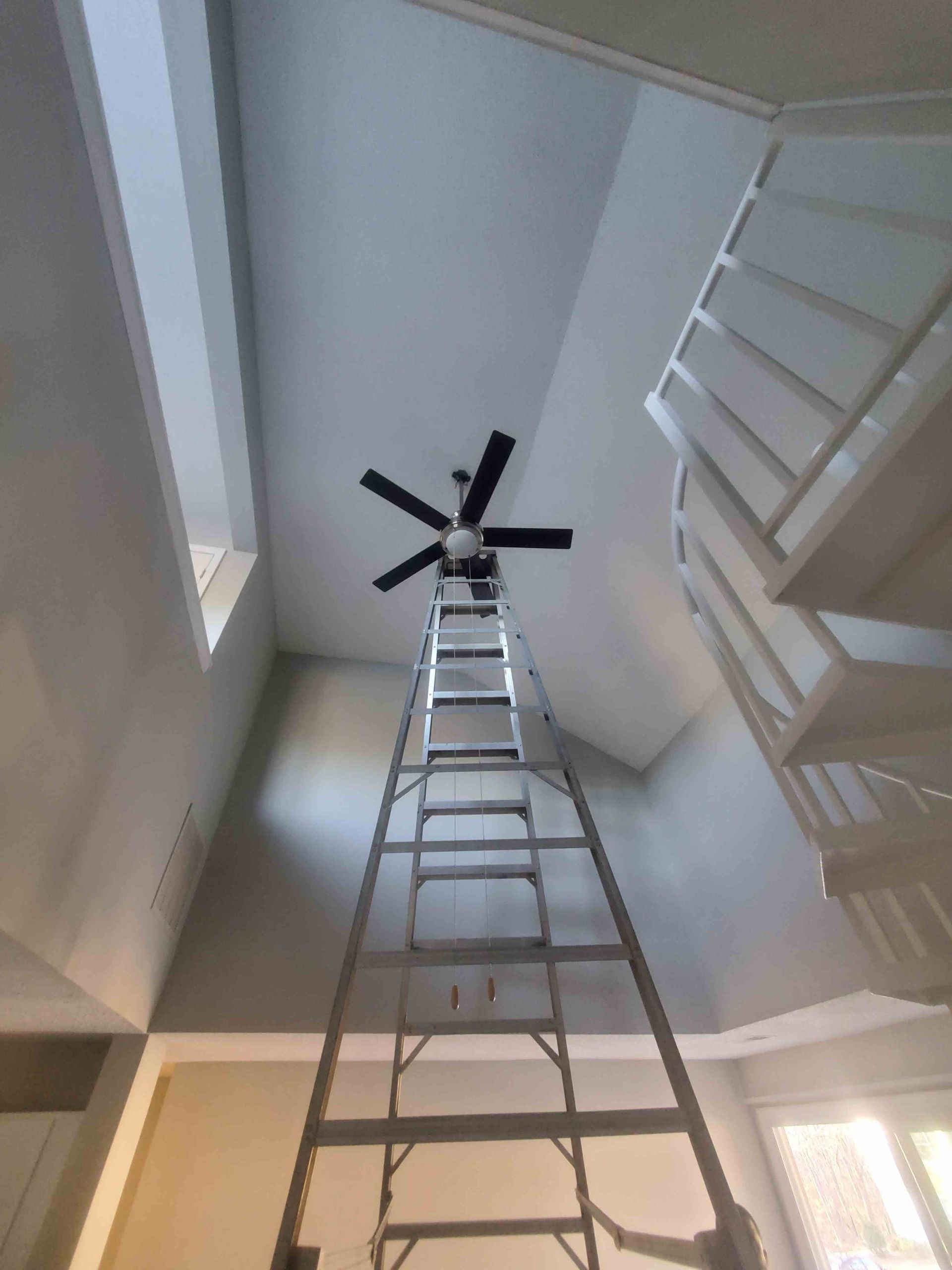 Tall ladder reaching a ceiling fan in a bright stairwell, viewed from below
