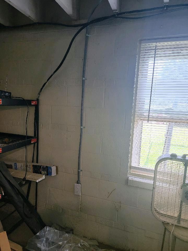 Dim basement room with cinderblock wall, window blinds, shelves, and a chair near hanging cables.