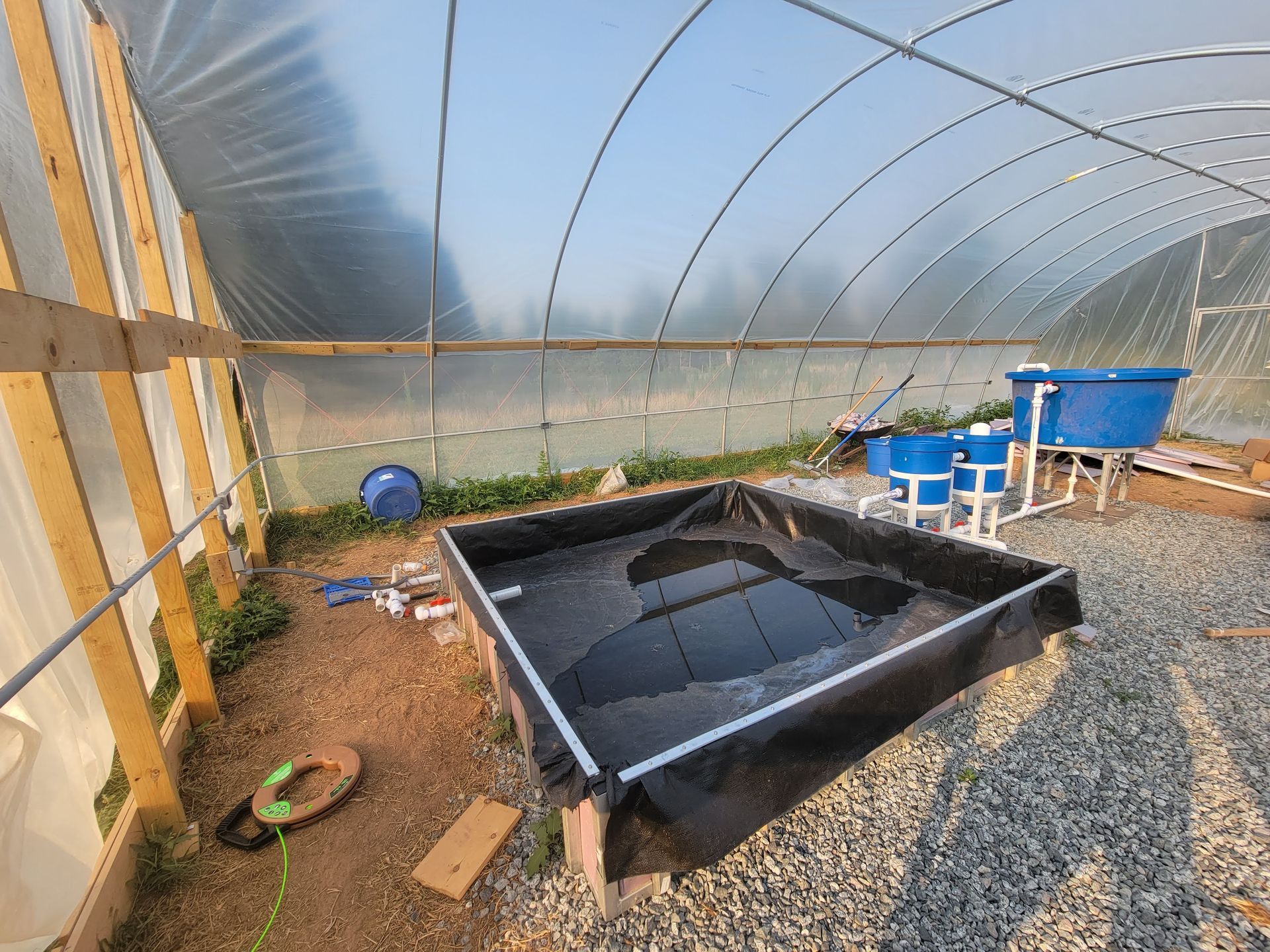 Black rectangular tank in a gravel greenhouse, with blue buckets and hoses nearby.
