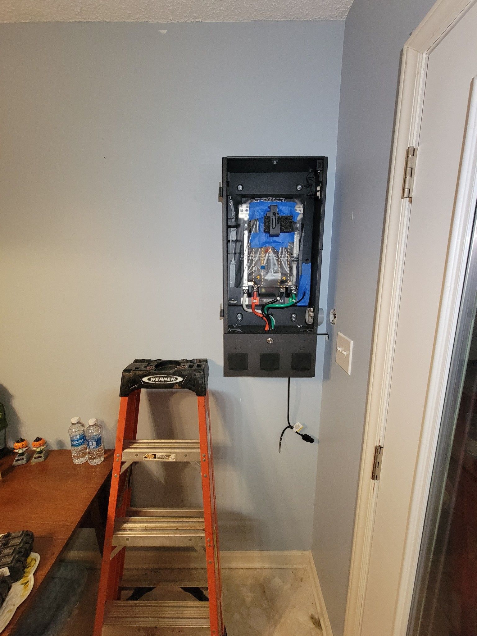 Open electrical panel in a room with a step ladder and table against a gray wall.